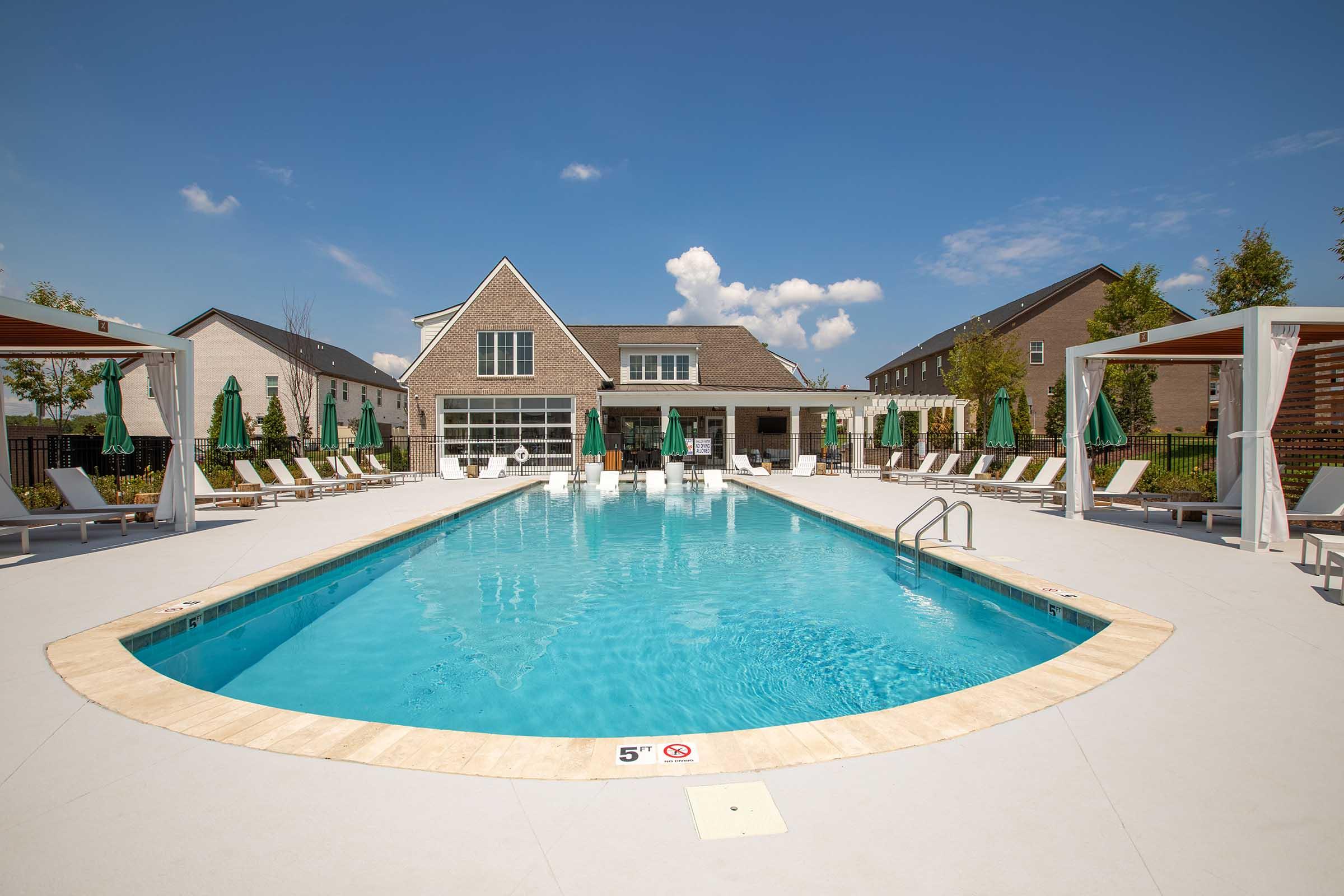 A sunny day at a modern outdoor swimming pool surrounded by lounge chairs and green umbrellas. The pool features a clear, inviting blue water and a depth marker. In the background, there are contemporary buildings and a bright blue sky with a few clouds.