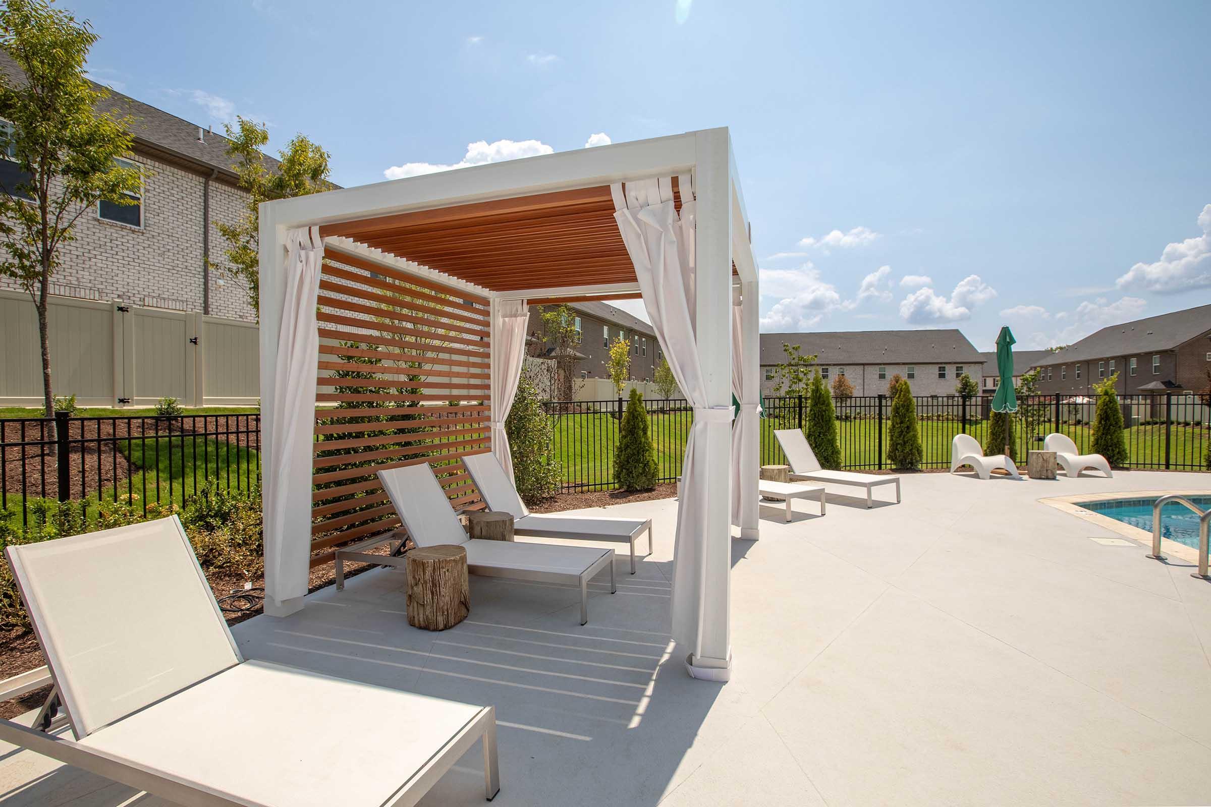 A sunlit poolside area featuring a shaded cabana with curtains, two lounge chairs, and a wooden stump table. The background includes green grass and a fenced area with several trees, along with a glimpse of residential buildings under a clear blue sky.