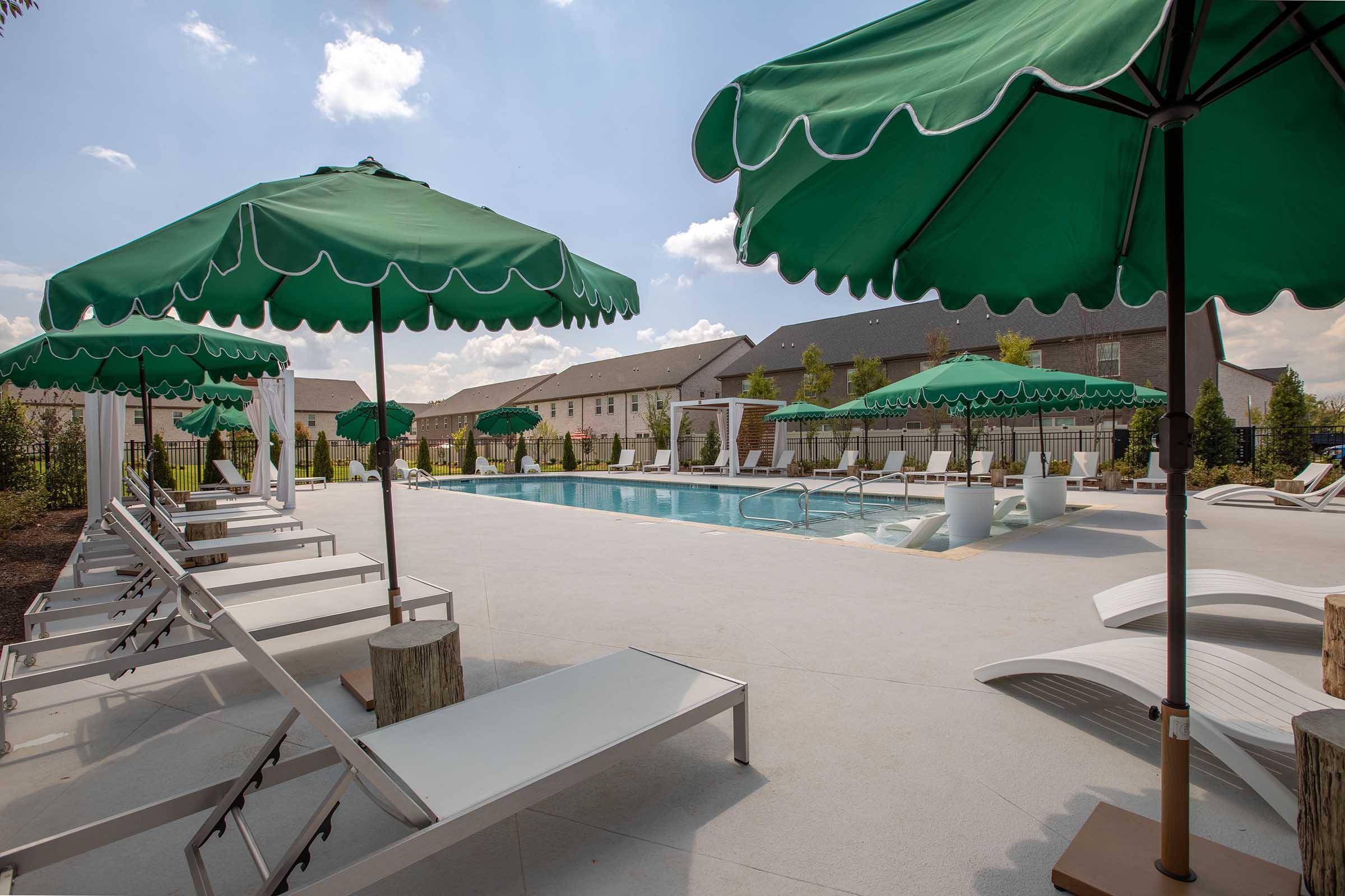 A serene swimming pool surrounded by lounge chairs and green umbrellas, set in a landscaped area with modern buildings in the background. The pool area features white decking and is bathed in sunlight under a clear blue sky, creating a relaxing atmosphere.