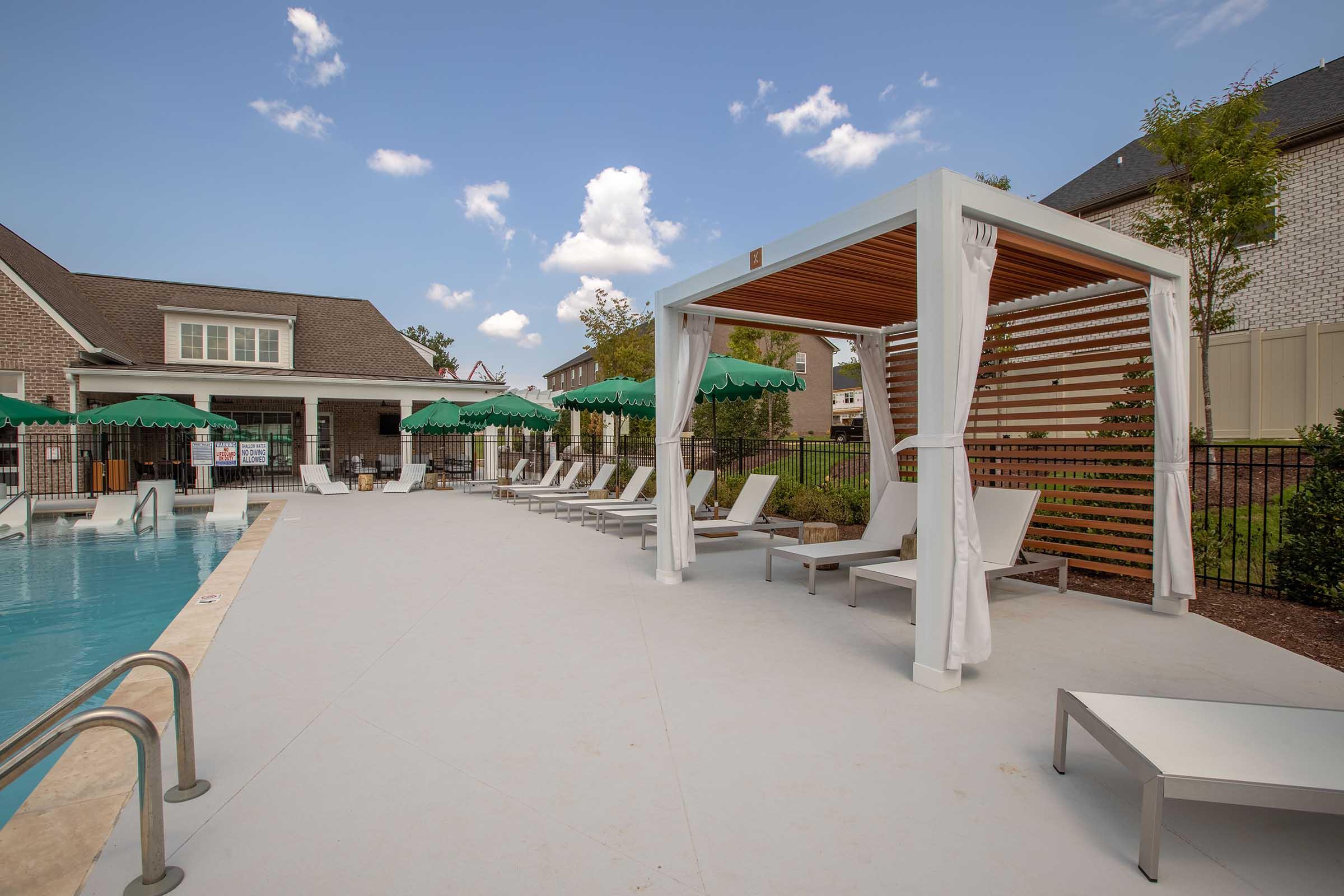 A serene pool area featuring a tranquil swimming pool, white lounge chairs, and a stylish cabana with draped curtains. Green umbrellas provide shade, surrounded by well-manicured landscaping and a blue sky with fluffy clouds. The setting invites relaxation and leisure.