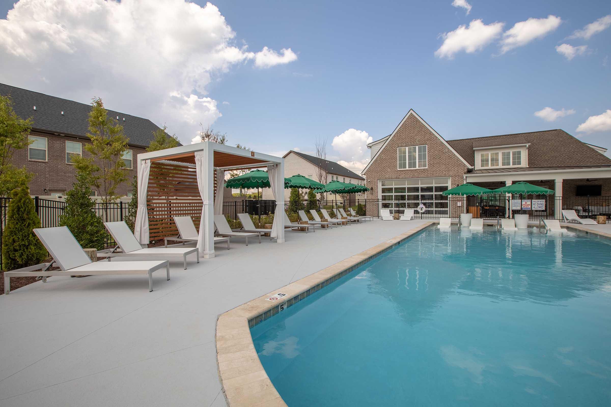 A spacious pool area featuring a sparkling blue pool surrounded by lounge chairs and green umbrellas. Tucked in the background is a modern brick building with large windows, and the sky is partly cloudy, creating a bright and inviting atmosphere for relaxation.