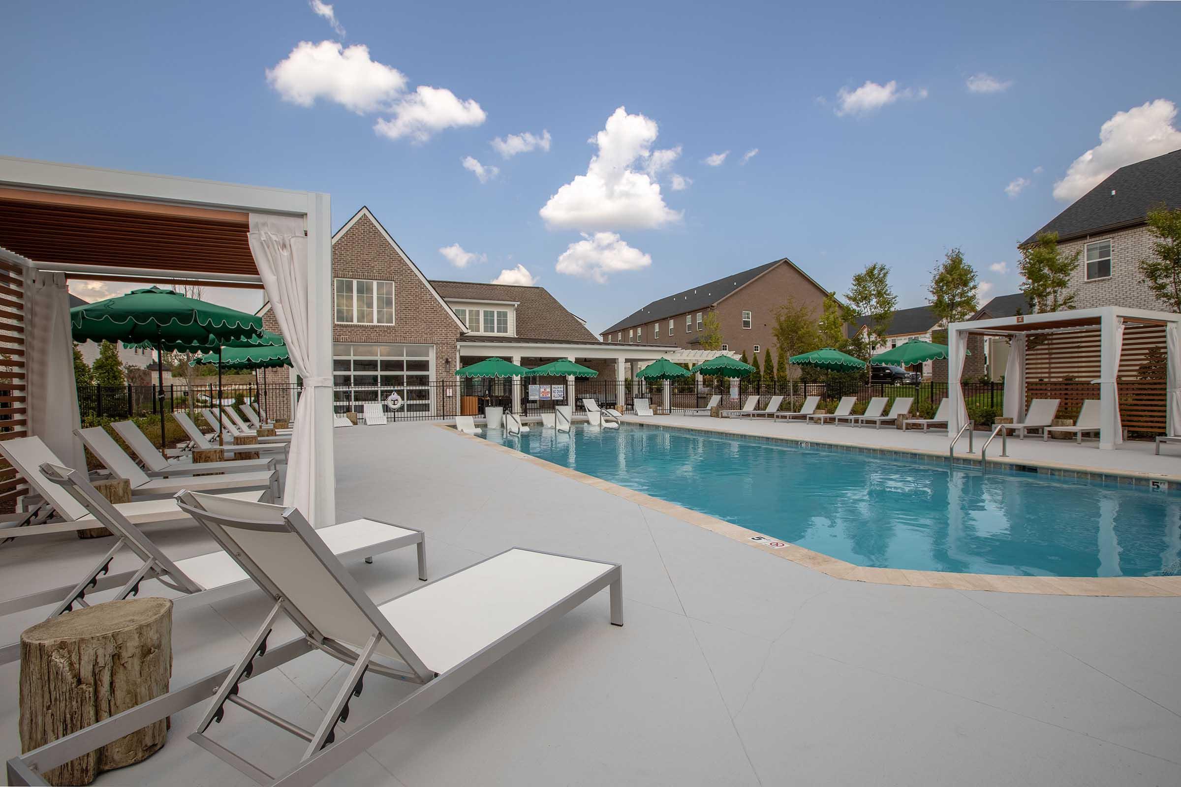 A spacious outdoor pool area featuring lounge chairs, white cabanas, and green umbrellas. The pool is surrounded by residential buildings under a clear blue sky with scattered clouds, creating a relaxing atmosphere for leisure and sunbathing.