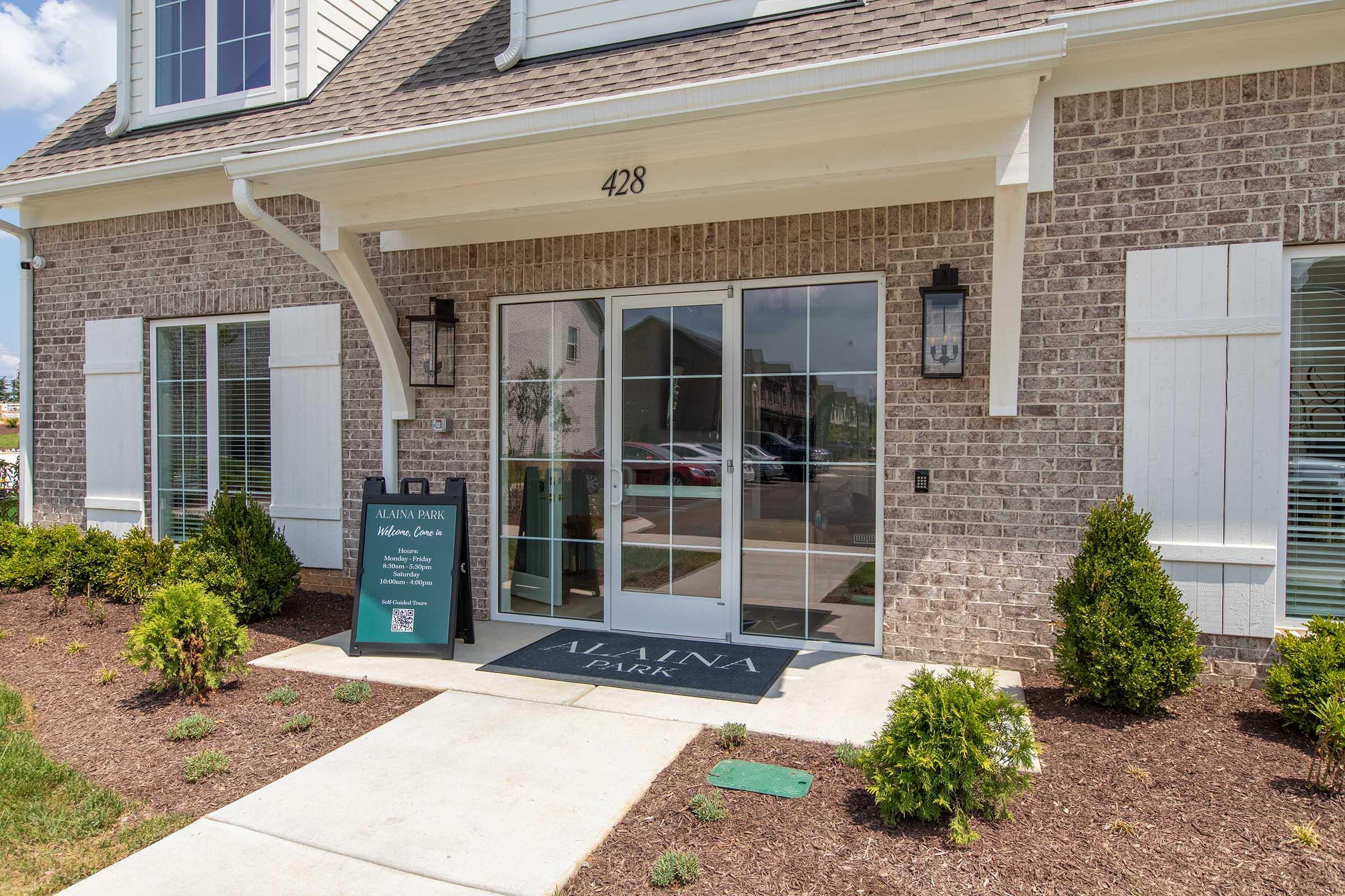 Entrance to the Alaina Park building at 428, featuring glass double doors with a welcome sign nearby. The area is landscaped with small shrubs and mulch. The exterior has a mix of brick and wood accents, and large windows are visible on either side of the entrance.