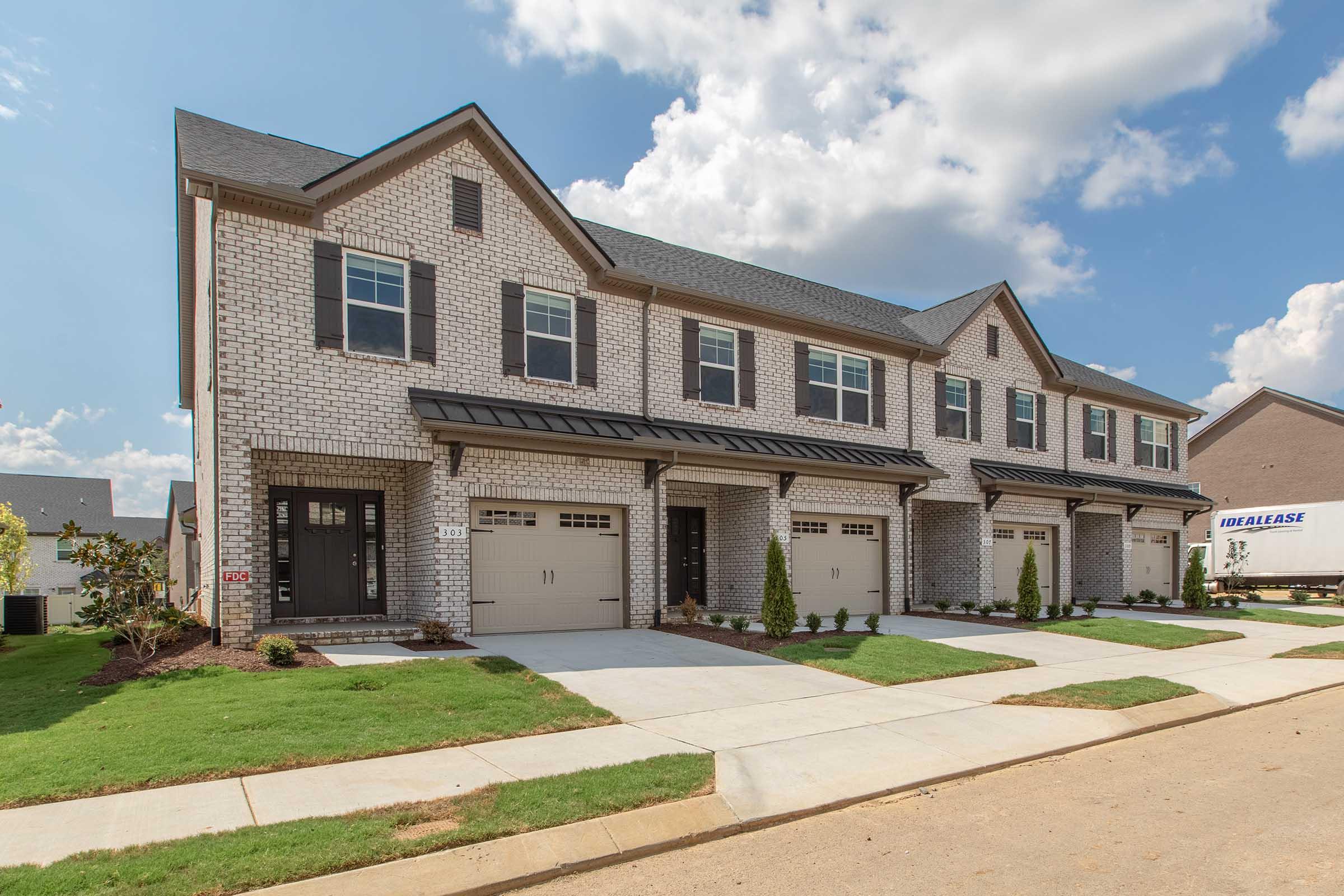 A newly constructed row of brick townhomes with attached garages and manicured lawns. The buildings feature gabled roofs and large windows under a clear blue sky, with a truck for leasing parked nearby. The setting appears suburban and well-maintained.