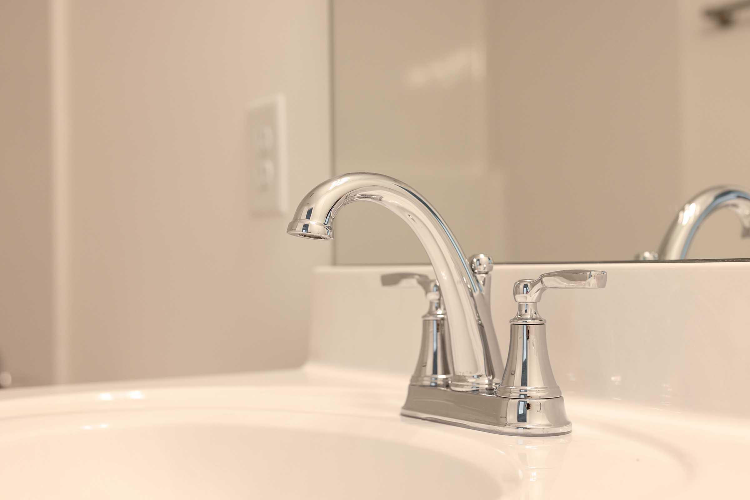 A close-up view of a modern chrome faucet on a white sink, with a mirror in the background. The faucet features elegant design elements, and the setting appears clean and minimalistic, typical of a contemporary bathroom. Soft lighting enhances the polished surface of the faucet.