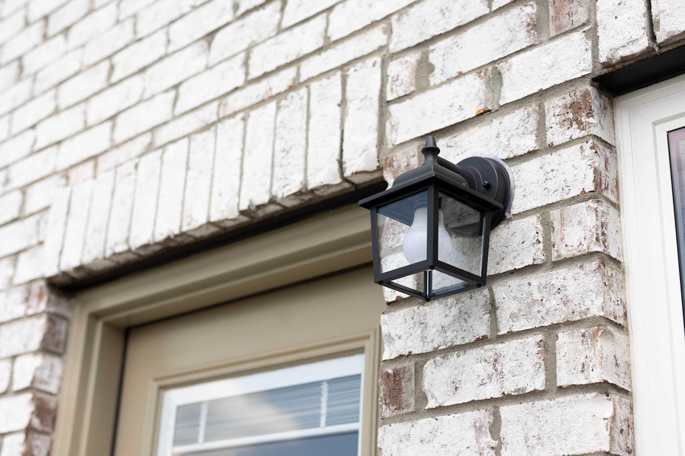 Wall-mounted lantern light fixture attached to a textured white brick exterior, situated near a door. The lantern has a classic design with a lantern shape and glass panels, housing a bulb inside. The door frame is neutral in color, complementing the brick wall.