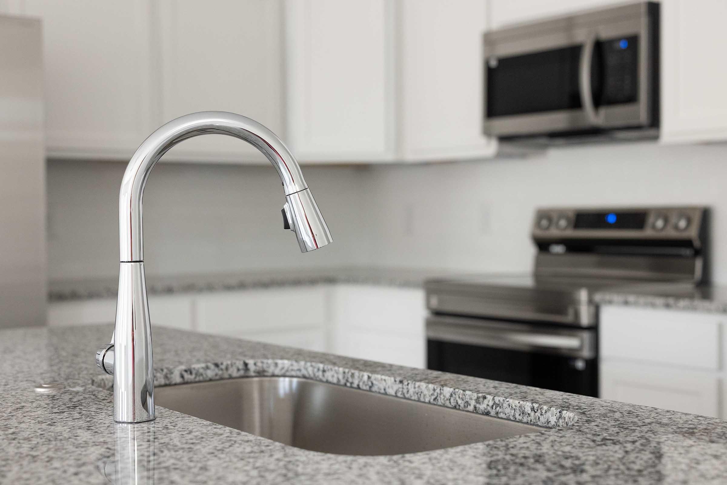 A modern kitchen featuring a stylish chrome faucet above a stainless steel sink, set on a granite countertop. In the background, there is a stainless steel microwave and stove, highlighting a sleek and contemporary design. The cabinetry is white, creating a bright and clean aesthetic.