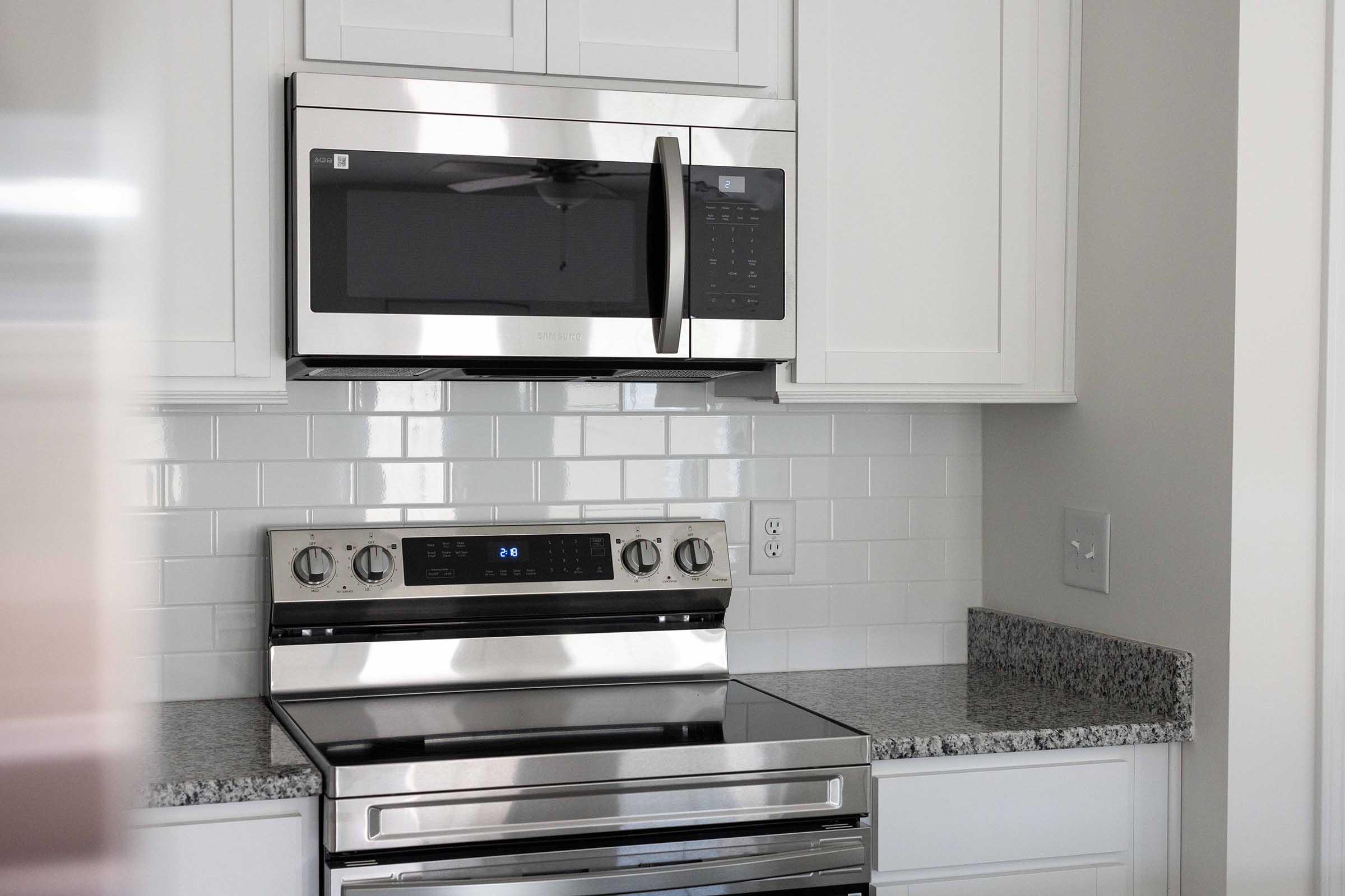 A modern kitchen featuring stainless steel appliances, including a microwave mounted above a sleek oven. The countertop is made of gray granite, and the backsplash consists of white subway tiles. The cabinetry is white, contributing to a clean and contemporary aesthetic.