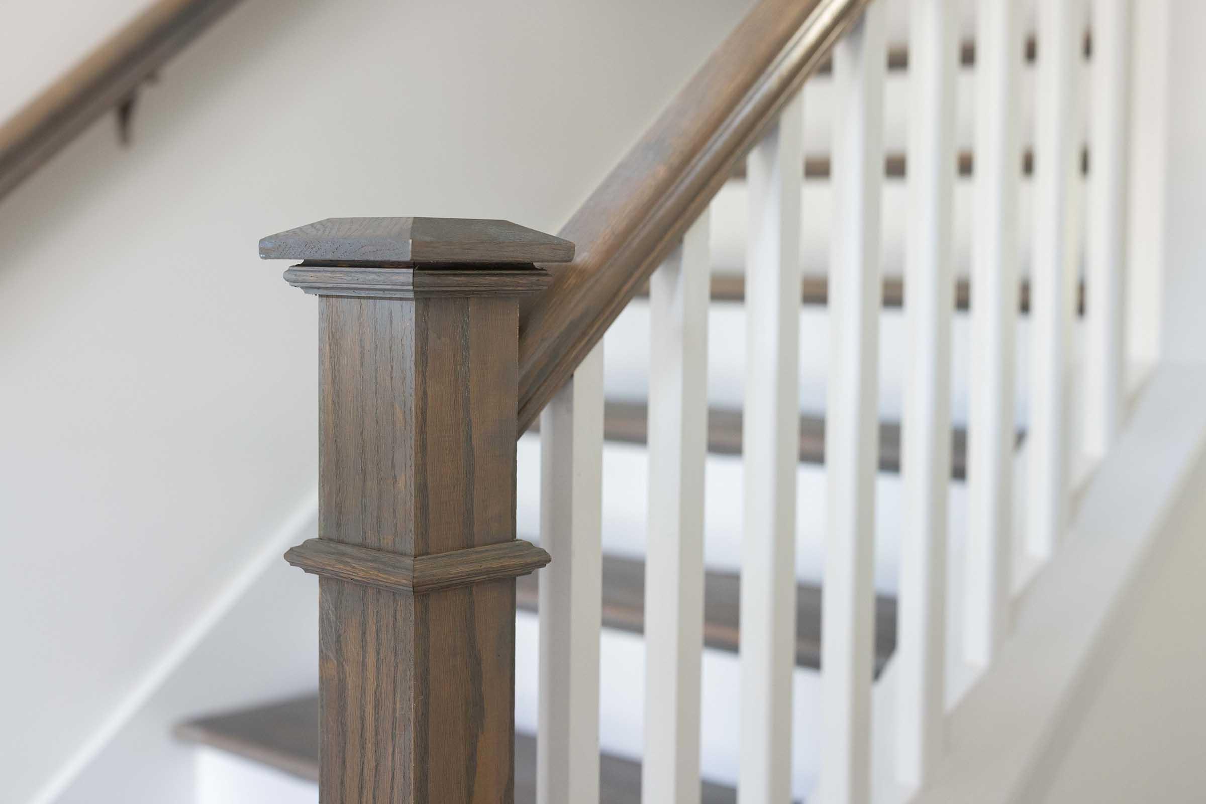 A wooden staircase with a polished handrail and a decorative newel post. The post features a hexagonal shape and a rich brown finish, while the railing is paired with white vertical balusters. The steps are simple and light-colored, creating a bright and modern look in the interior space.