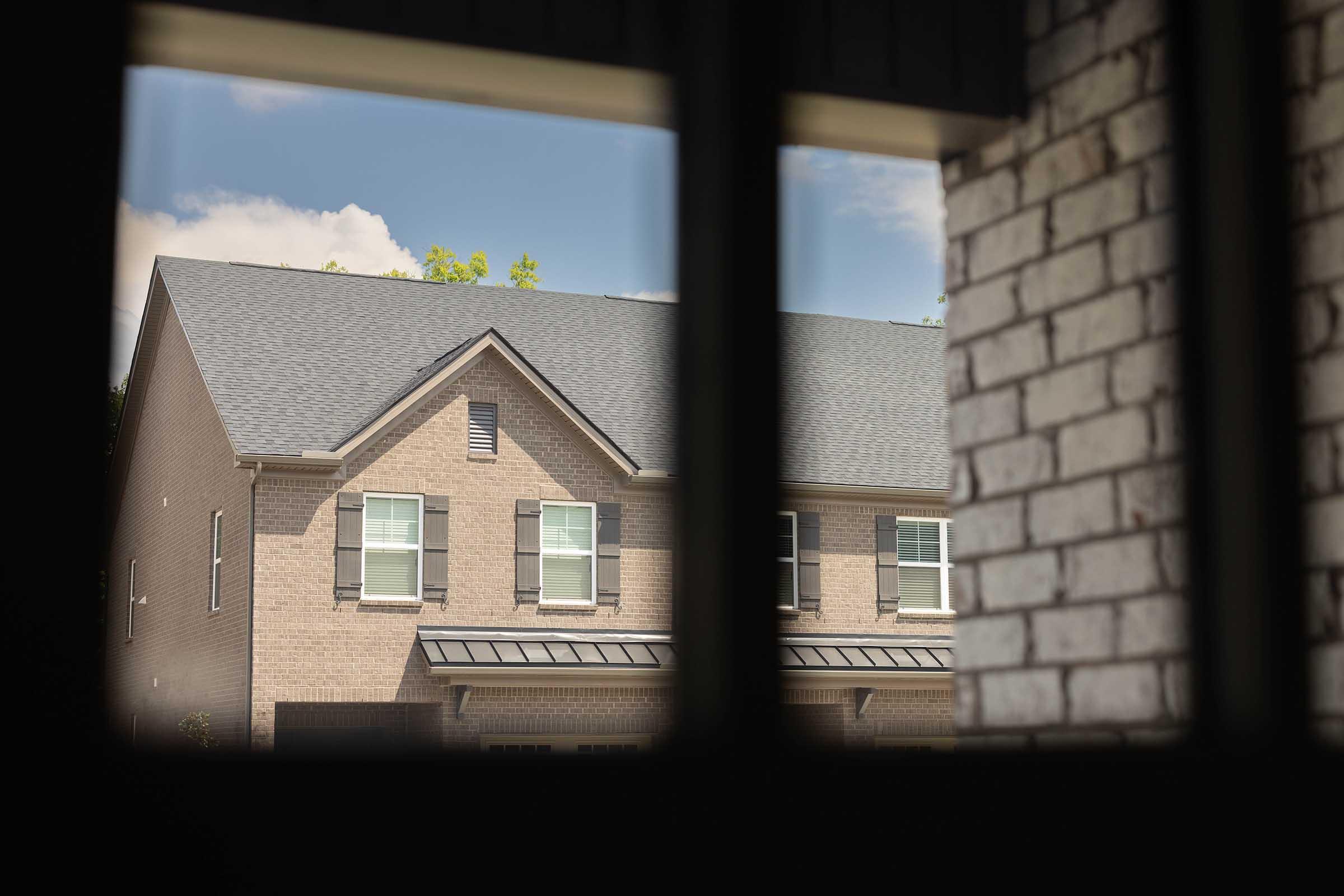 View of a residential building with a brick exterior and multiple windows. The roof is sloped with shingles, and there are green trees in the background against a clear blue sky. The perspective is from inside a structure, looking through a window frame.