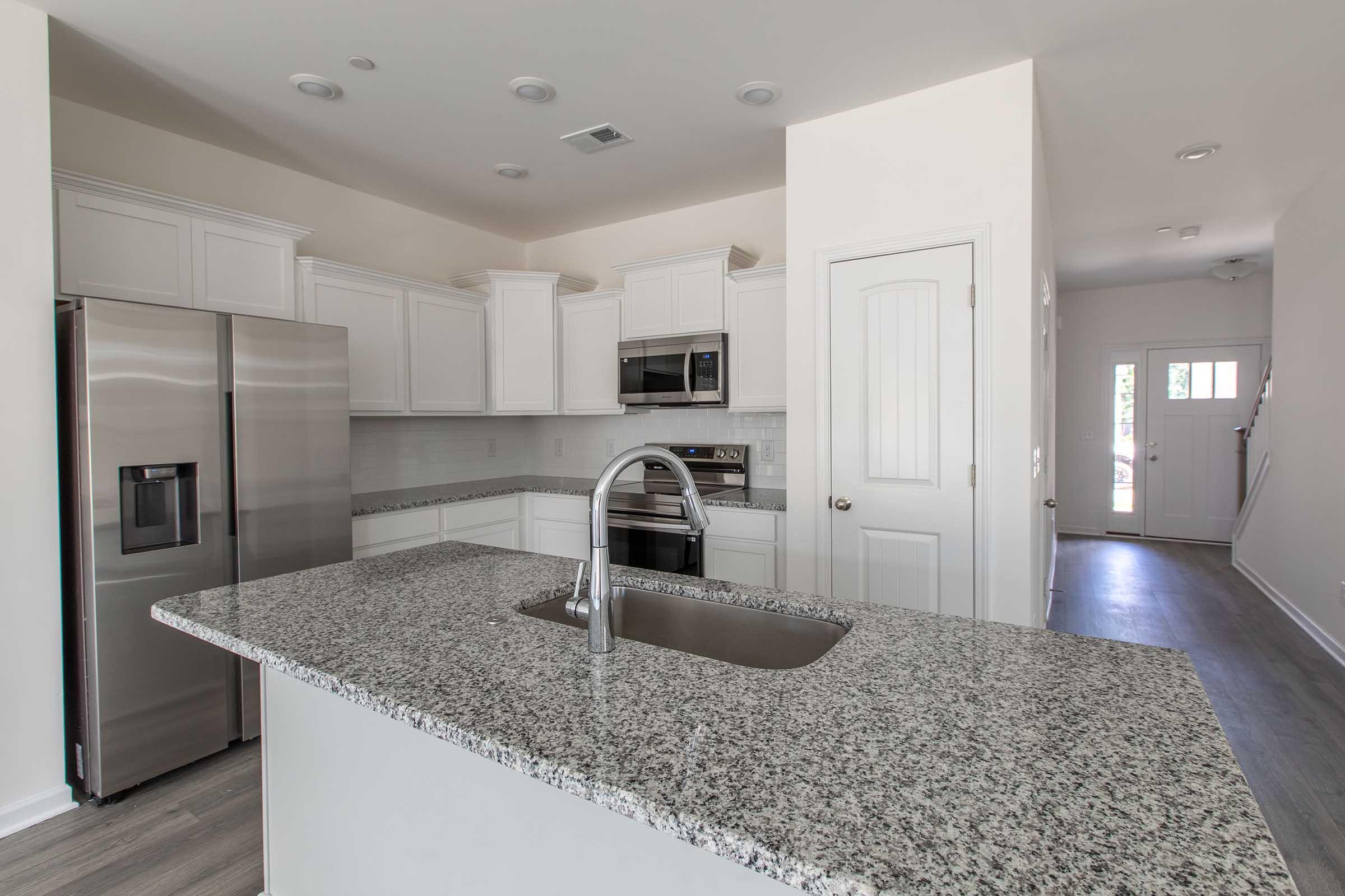 Modern kitchen featuring white cabinetry, granite countertops, stainless steel appliances including a refrigerator, microwave, and oven. The kitchen island has a sink and a sleek design, with a door leading to an outdoor area visible in the background. Bright, open layout with natural light.