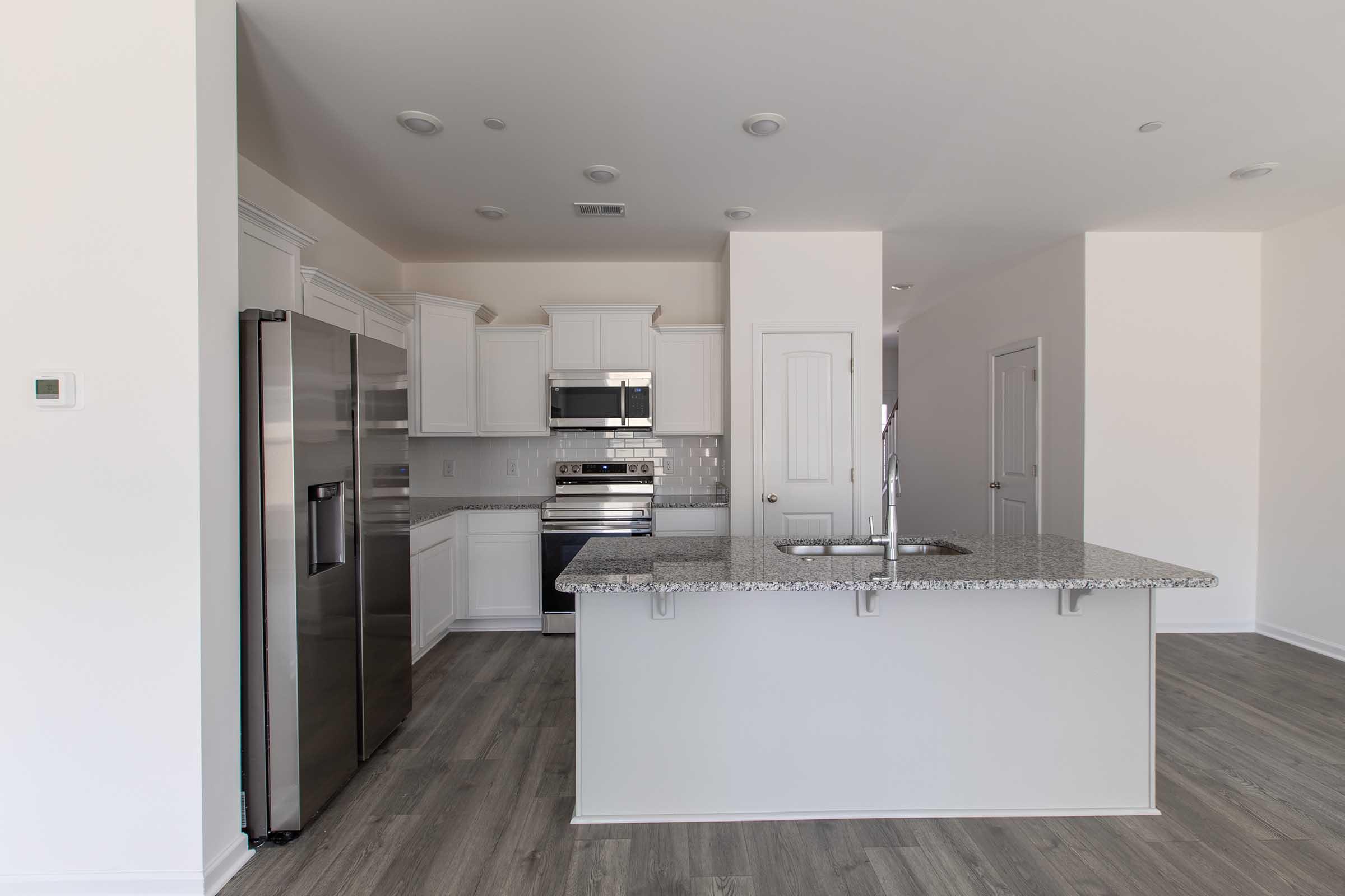 A modern kitchen featuring white cabinets, stainless steel appliances, and a large granite island. The open layout includes tile flooring and neutral-colored walls, withample natural light illuminating the space. A pantry door is visible, along with another door leading to a separate area.