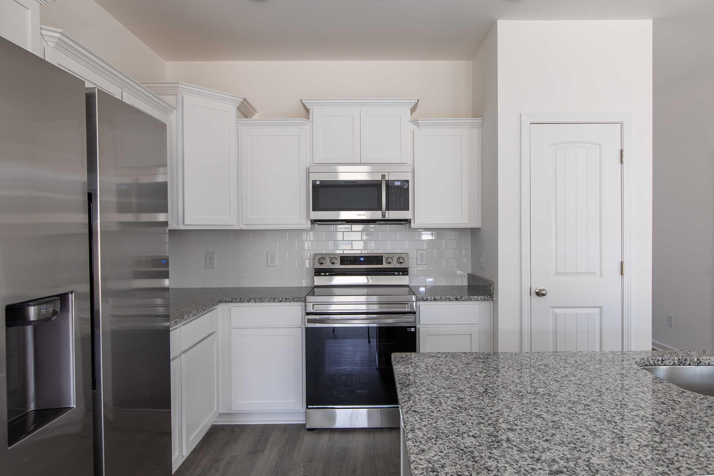 Modern kitchen featuring white cabinetry, stainless steel appliances including an oven and microwave, and a granite countertop. The space is well-lit, with a door leading to another area, and a sleek, minimalist design.