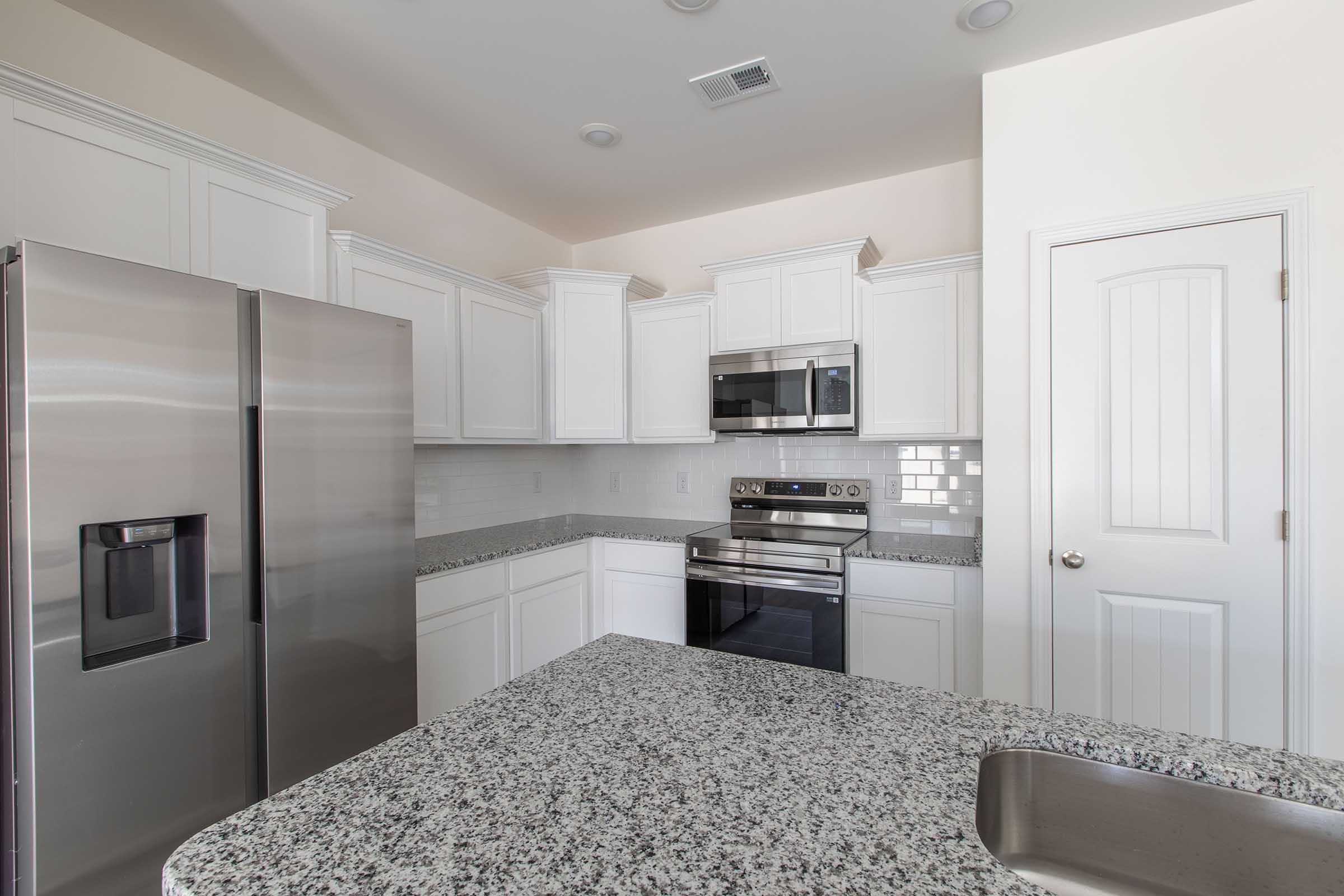 Modern kitchen featuring white cabinetry, stainless steel appliances including a refrigerator and oven, and a granite countertop. The space is bright with natural light, showcasing a clean and contemporary design.