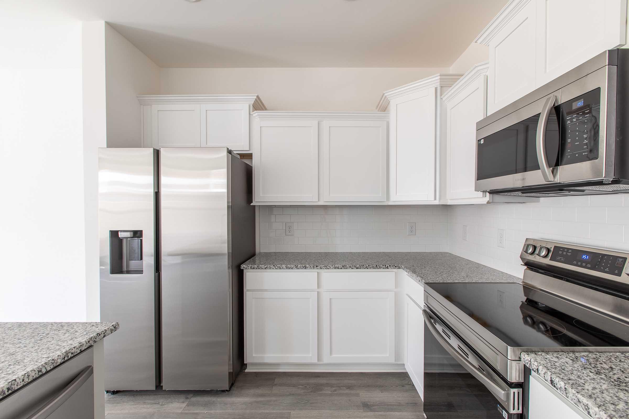 Modern kitchen interior featuring white cabinetry, stainless steel refrigerator, microwave, and oven. The countertops are gray with a speckled pattern, and the flooring is light-colored. The overall design is sleek and contemporary, emphasizing functionality and style.