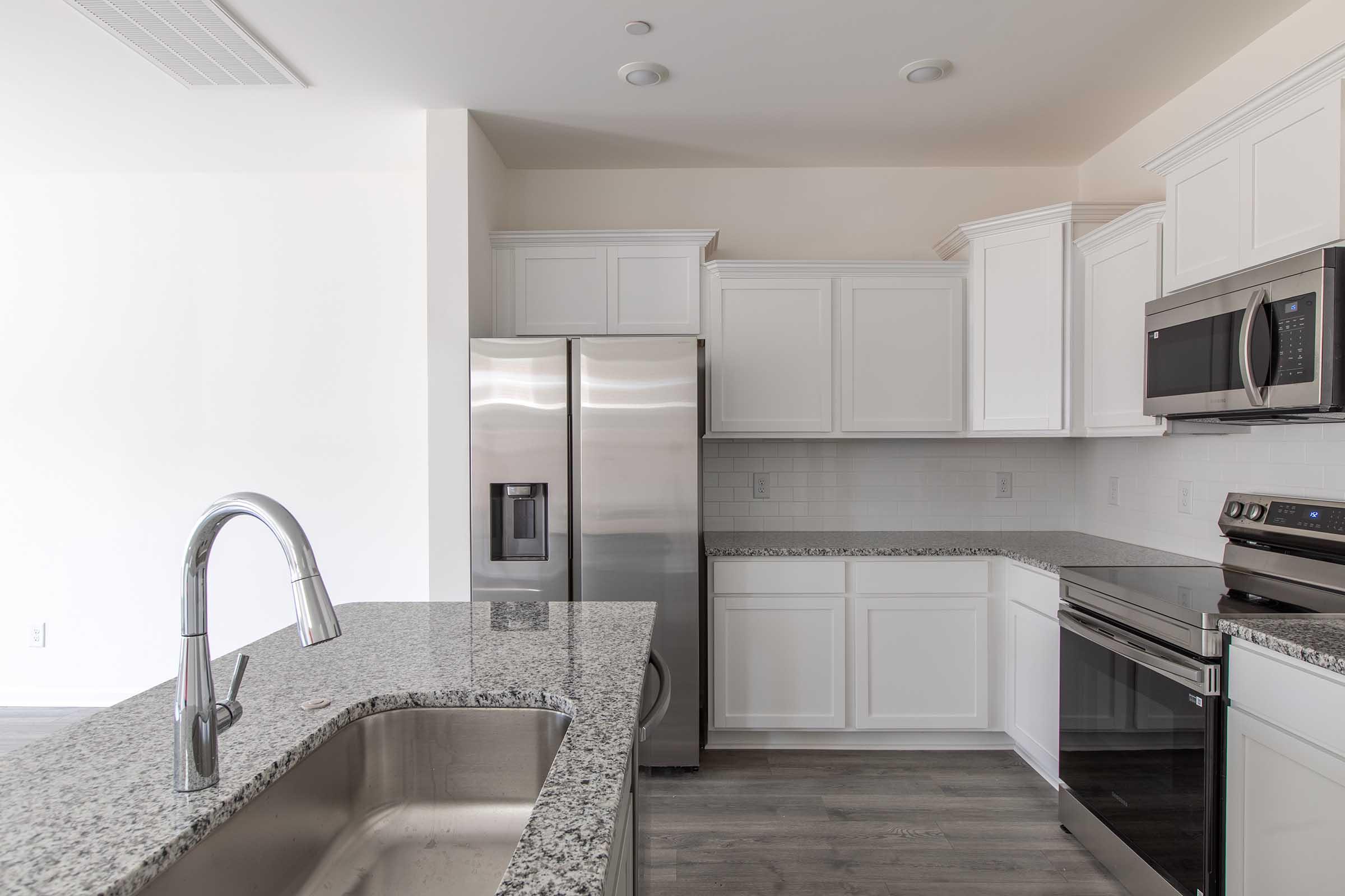Modern kitchen featuring white cabinetry, stainless steel appliances including a refrigerator and oven, and a granite countertop with a double sink. The space is well-lit and has a clean aesthetic with a light color scheme and wooden flooring.