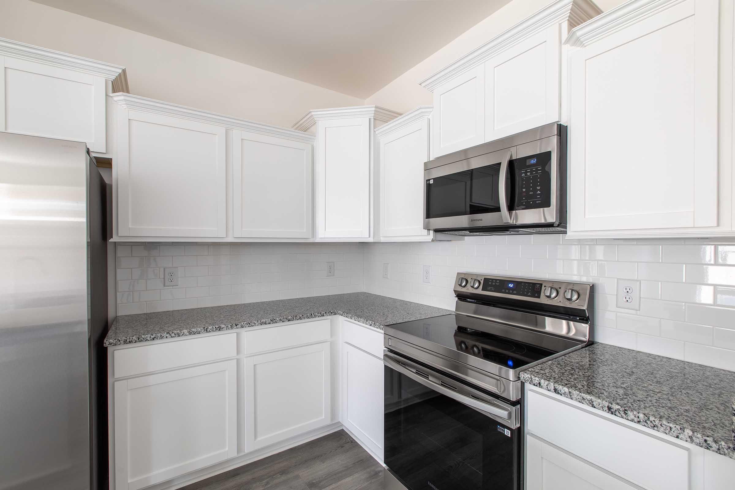 Modern kitchen featuring white cabinetry, stainless steel appliances including a microwave and oven, and a granite countertop. The space has a clean design with a tiled backsplash and wood-like flooring, creating a bright and inviting atmosphere.