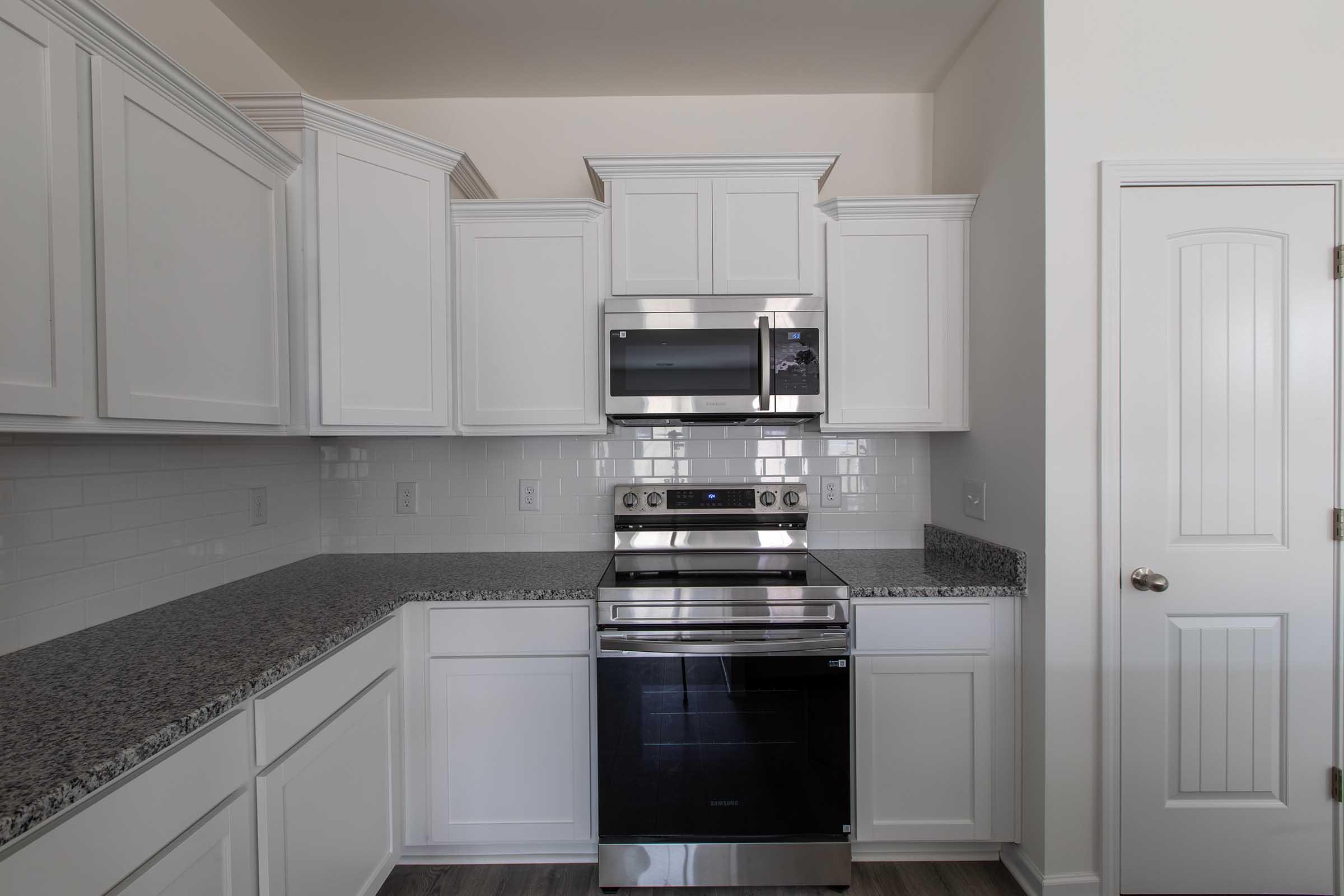 A clean, modern kitchen featuring white cabinetry, a stainless steel microwave, and an oven range. The countertops are made of granite, and there’s a white pantry door visible on the right. The overall design is bright and contemporary, emphasizing a spacious and organized layout.