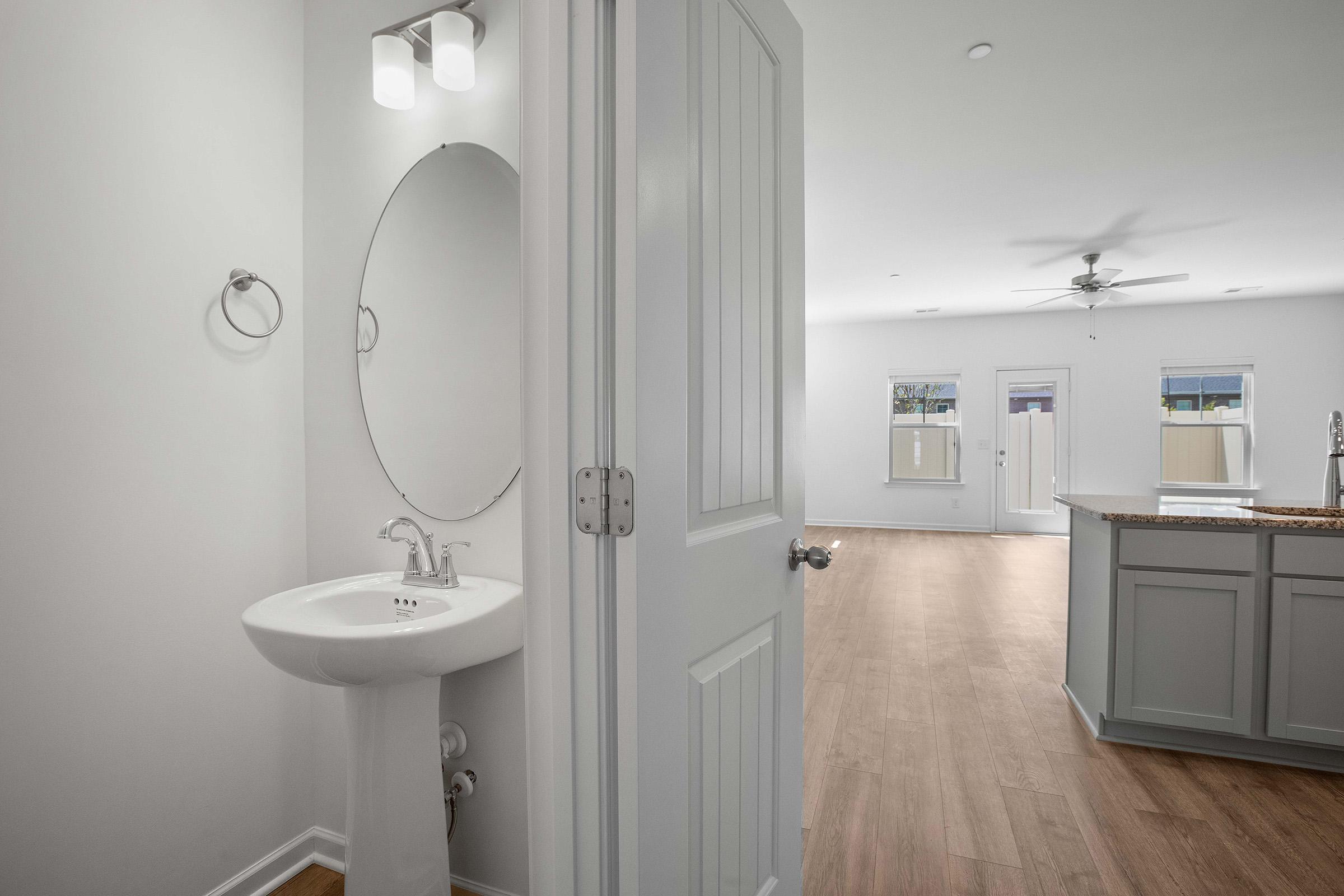 A glimpse of a modern interior featuring a small half-bath with a round mirror and sink on the left, while the right side shows a spacious room with light hardwood flooring, a ceiling fan, and access to outdoor areas through glass doors.