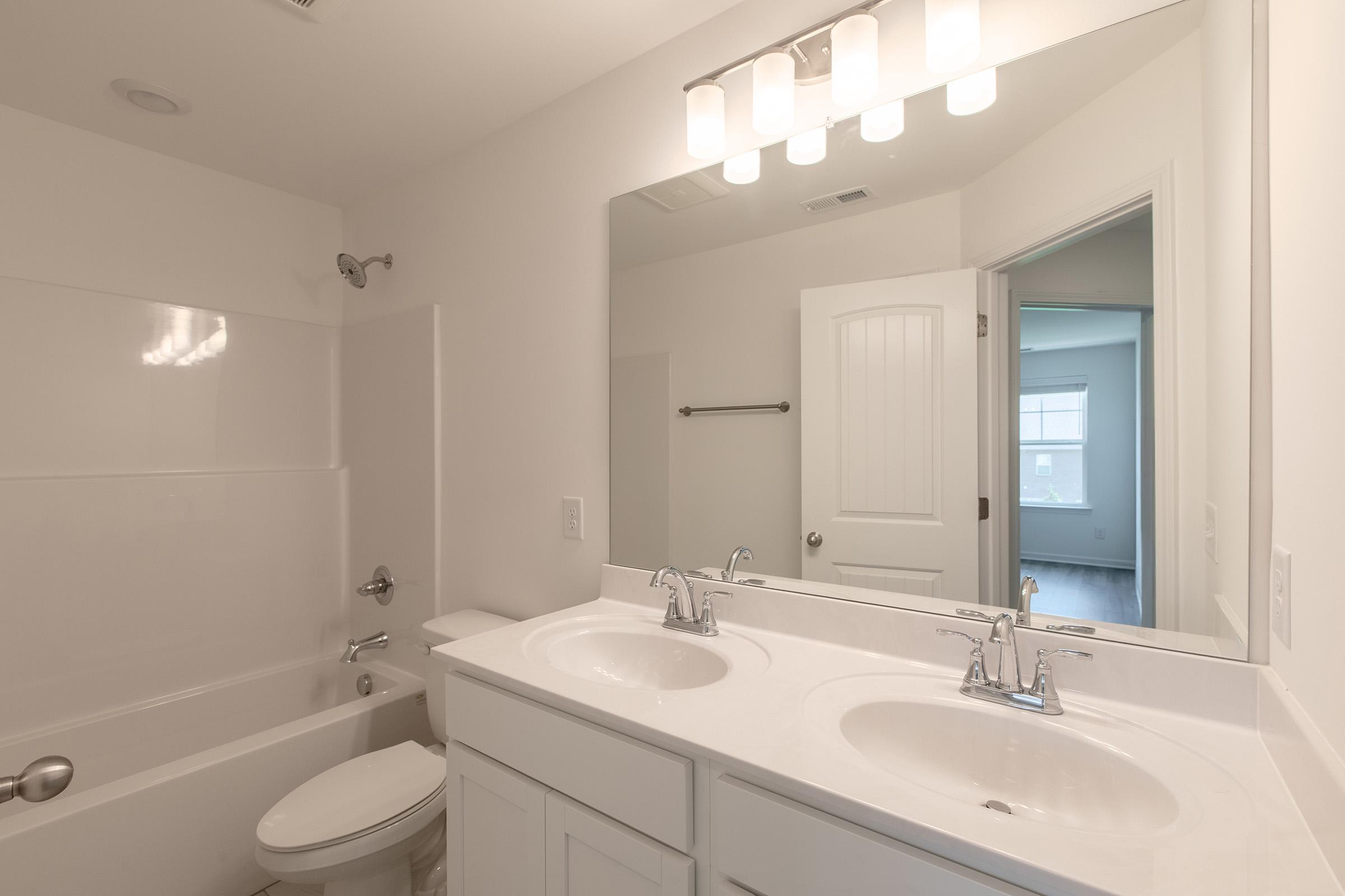 A modern bathroom featuring a double vanity with two sinks, a large mirror above, and a bathtub with a shower. The walls are painted white, and there's a minimalist design. A door leads to another room, and natural light enters through a window in the adjacent space.