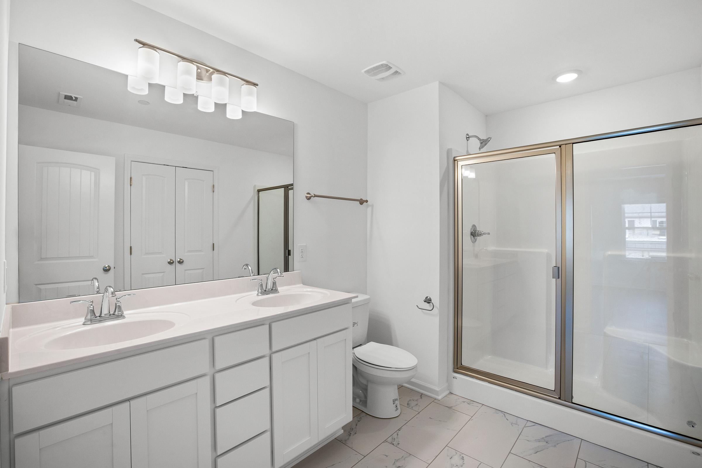 A modern bathroom featuring a double vanity with two sinks, a large mirror above, a shower area with glass doors, a toilet, and white cabinetry. The walls are painted white, and the floor has light-colored tiles. Natural light enters through a window, creating a bright and clean atmosphere.