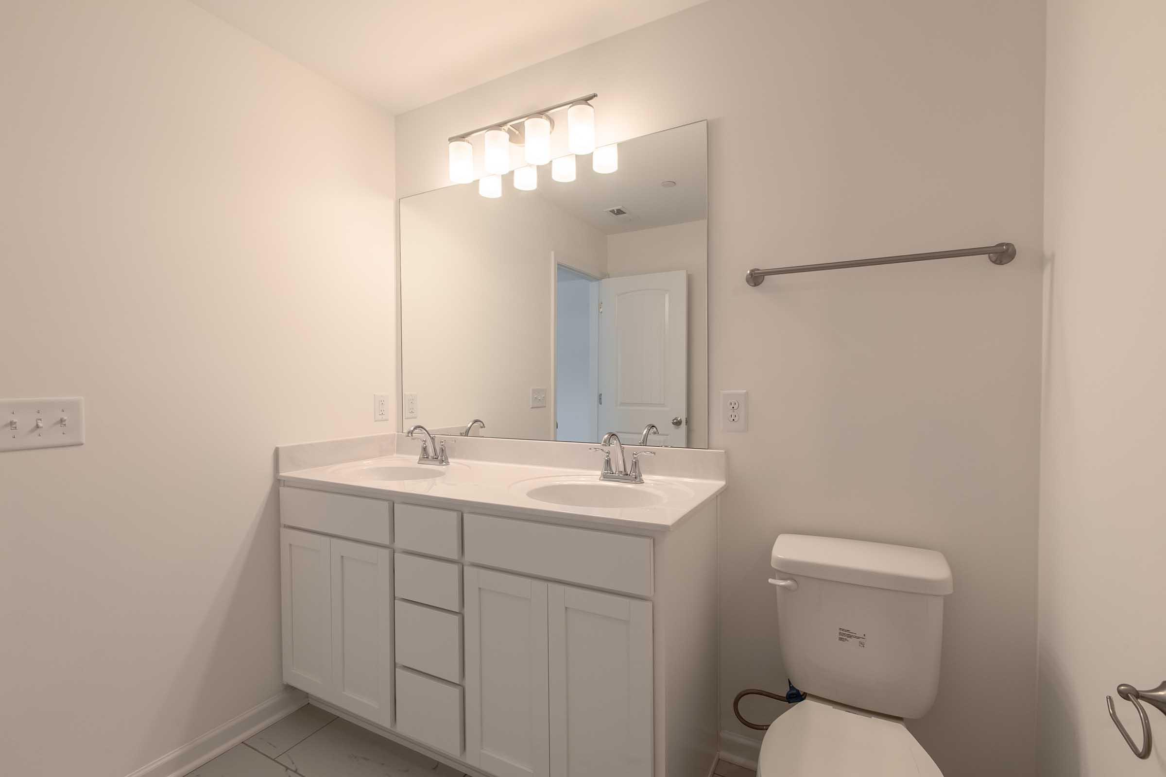 A modern bathroom featuring a double sink vanity with white cabinetry, a large mirror above, a wall-mounted towel bar, a toilet, and neutral-colored walls. The room is well-lit with overhead lighting and has a clean, minimalist aesthetic.