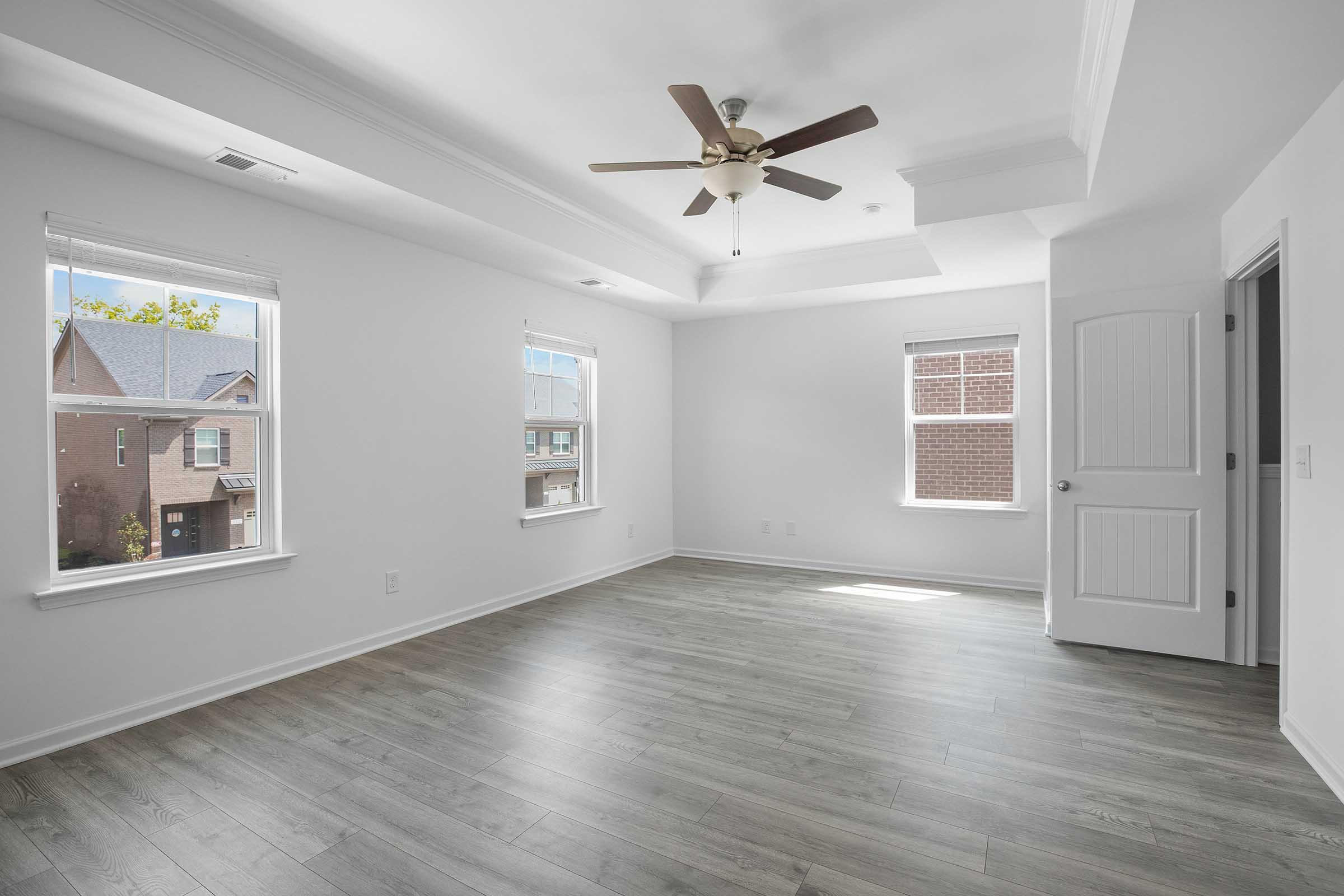 A spacious, empty room with white walls and a ceiling fan. Light streams through two windows, illuminating the hardwood floor. A door on the right leads to another room. The room is uncluttered, creating a bright and airy atmosphere.