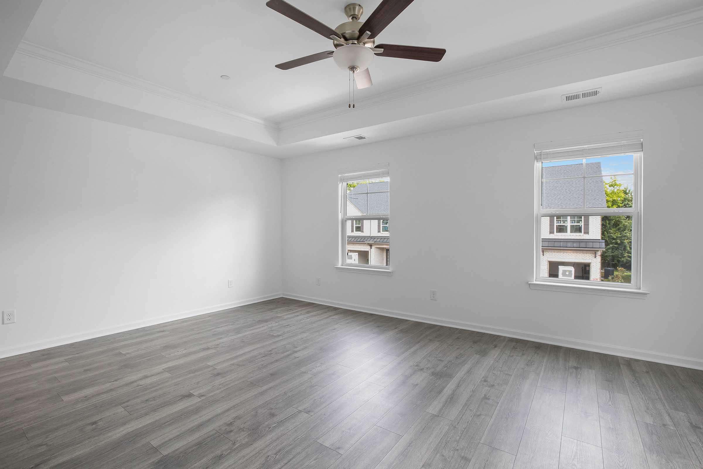 A spacious, empty room with light gray walls and a wooden floor. There are two windows allowing natural light, with a ceiling fan overhead. The room features a white tray ceiling, creating a bright and airy atmosphere, suitable for various design options.