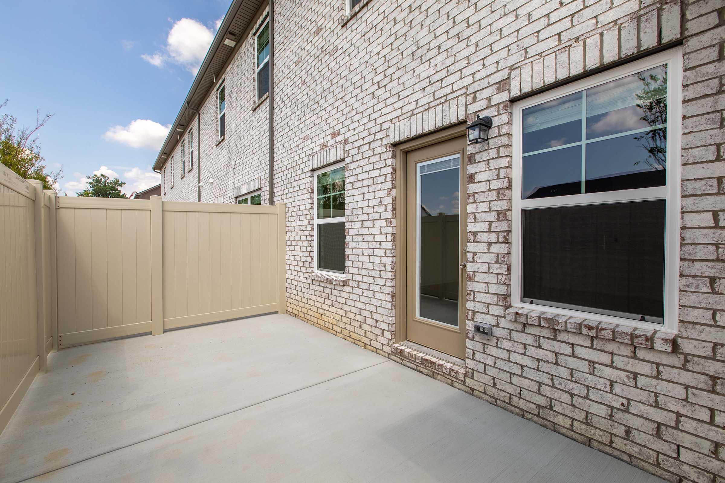 A view of the back of a brick house, featuring a concrete patio area with a tan privacy fence. There is a door leading inside and several windows allowing natural light, framed by a clear blue sky with a few clouds. The overall setting looks clean and well-maintained.