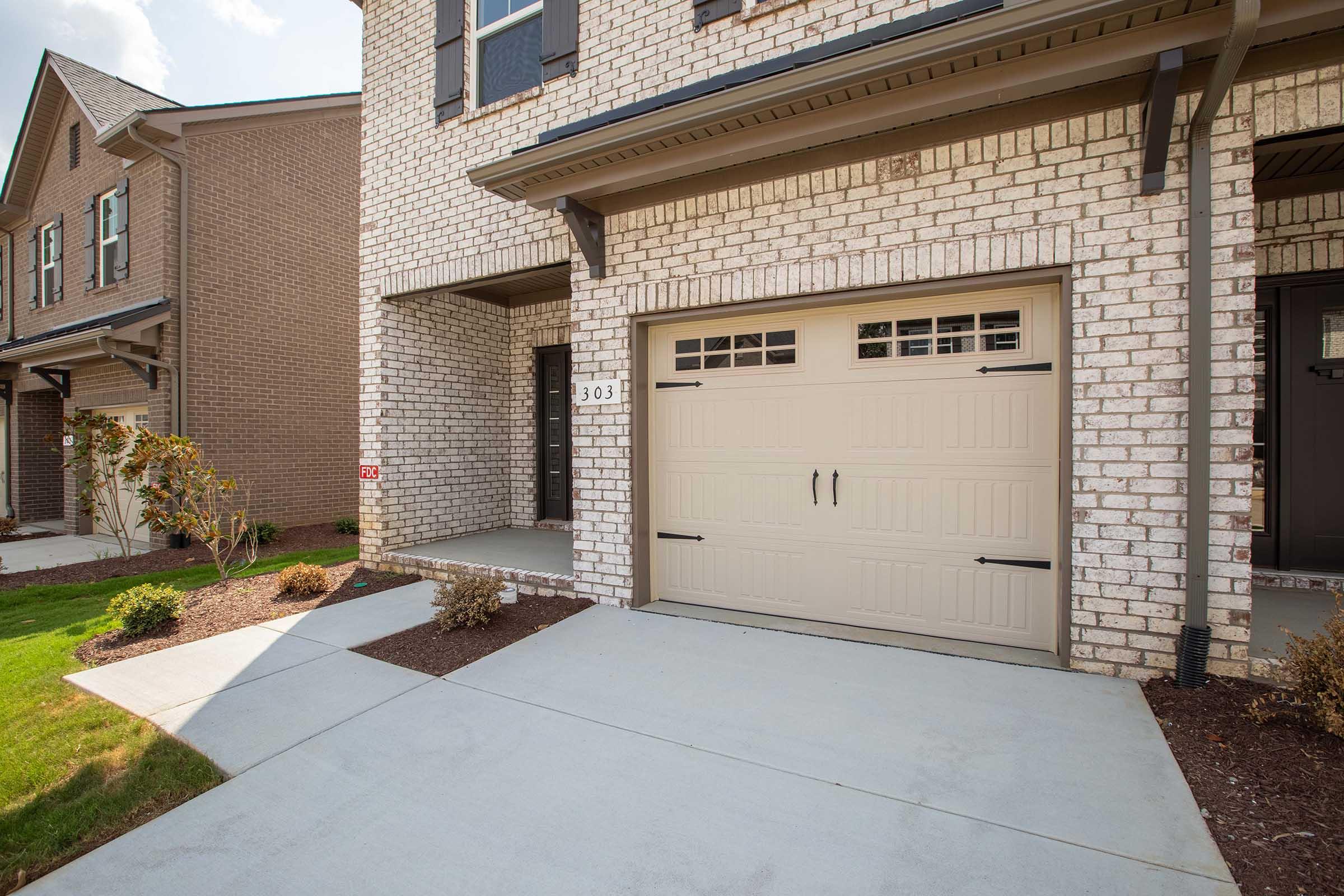 A residential building showcasing a brick exterior with a beige garage door, a concrete driveway, and manicured landscaping. The house features a front porch area and is situated in a neighborhood with similar homes. Bright blue skies add to the inviting atmosphere.