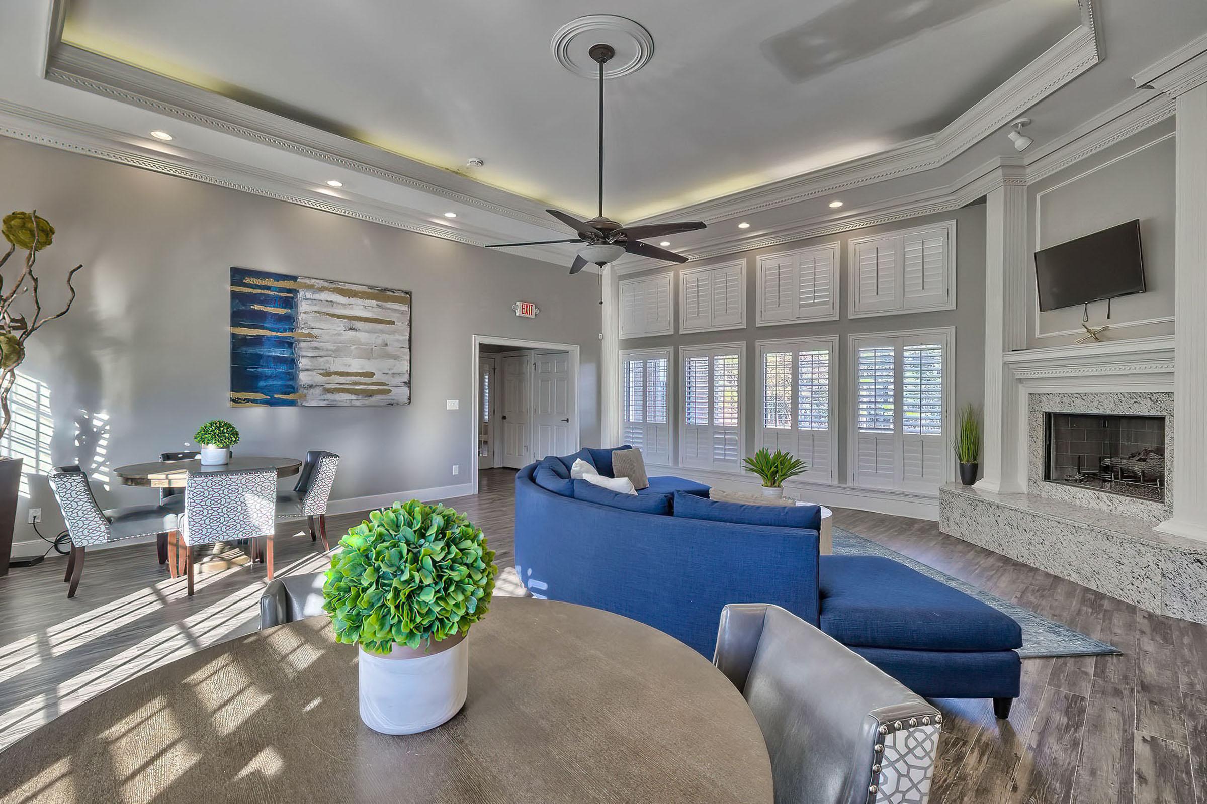 A bright and modern living room featuring a blue sectional sofa, a circular wooden table with four stylish chairs, and a decorative plant. The room is enhanced by large windows with shutters, hardwood flooring, and a cozy fireplace, creating a welcoming and spacious atmosphere.