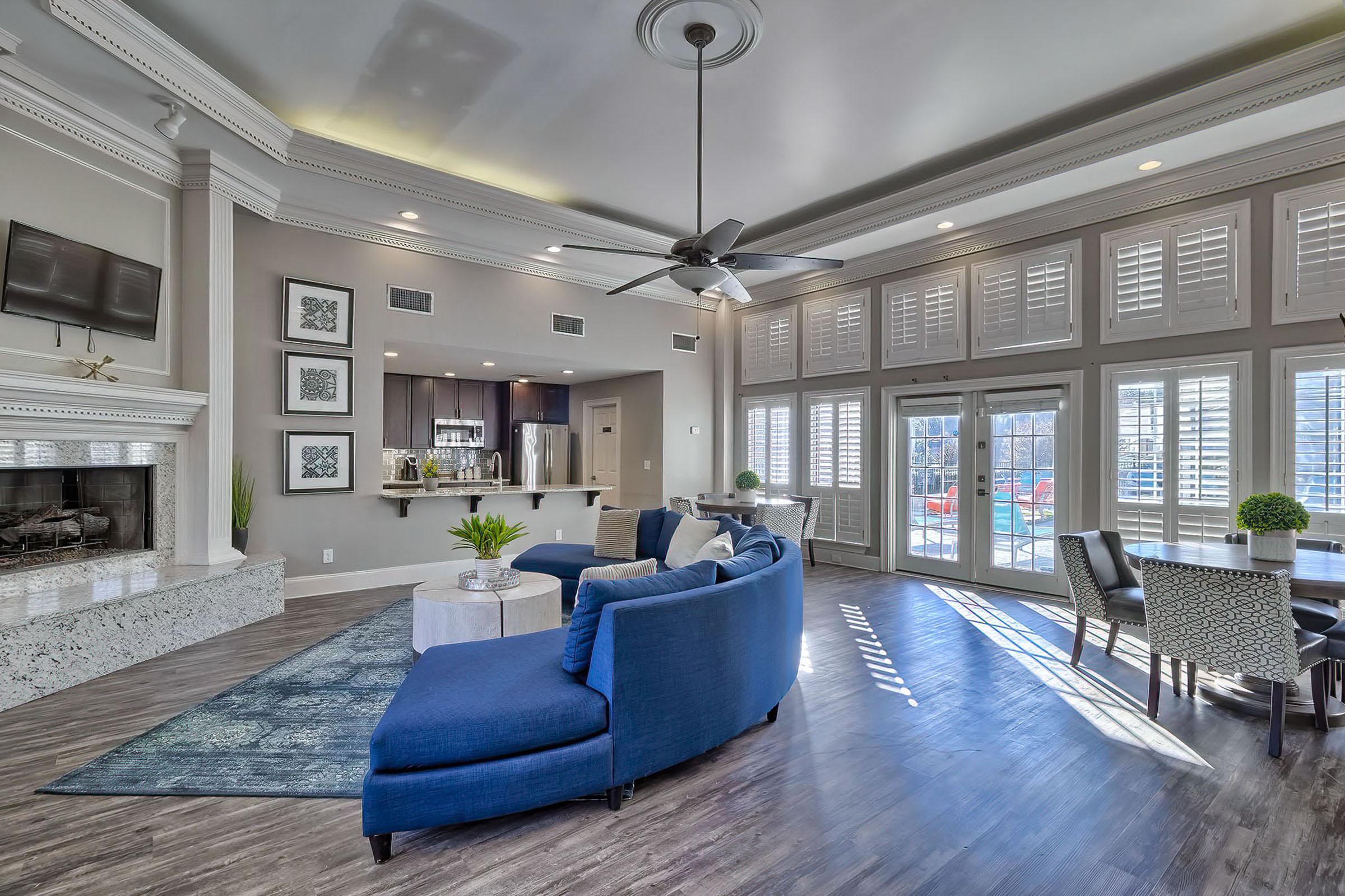 A spacious living room with a modern design featuring a blue sectional sofa, a round coffee table, and a textured area rug. Large windows with shutters allow natural light to fill the space, highlighting a stylish fireplace and a dining area nearby. The kitchen is partially visible in the background.