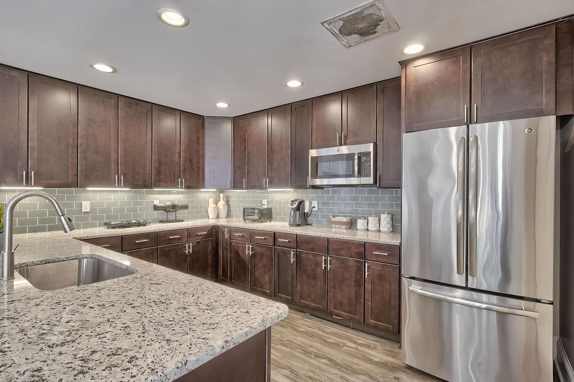 Modern kitchen featuring dark wood cabinets, a grey tiled backsplash, stainless steel appliances including a microwave and refrigerator, and a granite countertop with an undermount sink. Soft lighting illuminates the space, creating a warm and inviting atmosphere.