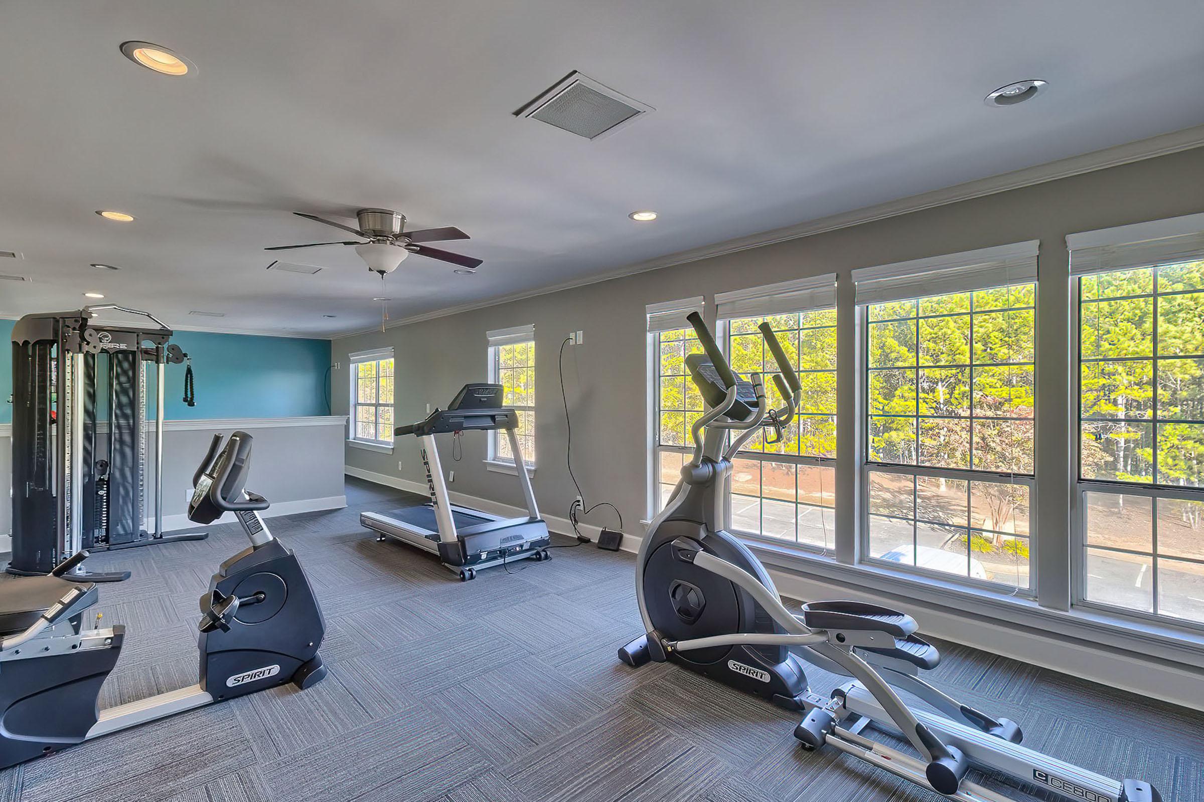 A well-lit fitness room featuring exercise equipment, including a treadmill, stationary bike, and elliptical machine. Large windows allow natural light to fill the space, showcasing a clean and inviting environment. The walls are painted in soft colors, adding to the modern aesthetic.