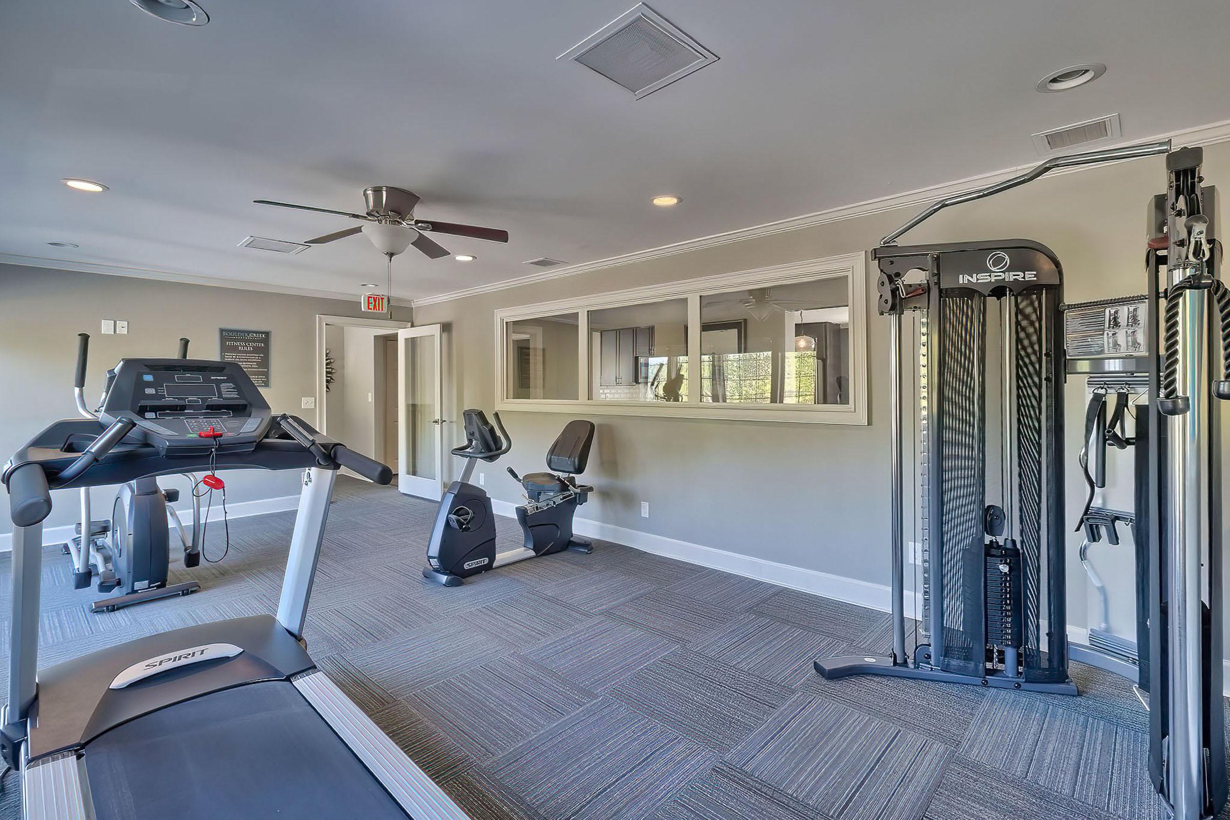 A modern fitness room featuring a treadmill, stationary bike, and multi-station gym equipment. The space is well-lit with ceiling lights and a ceiling fan, and has a neutral color scheme with carpeted flooring. Large windows provide natural light, creating an inviting atmosphere for exercise.