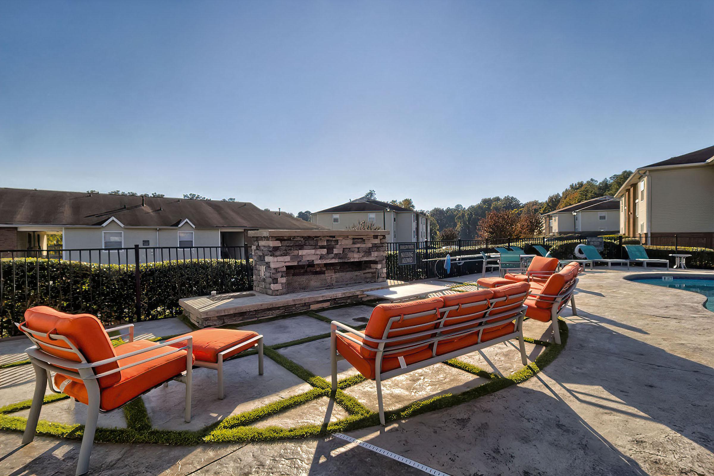 A serene outdoor area with bright orange lounge chairs surrounding a stone fireplace. In the background, there are apartment buildings and trees under a clear blue sky. The scene conveys a welcoming space for relaxation and social gatherings by a swimming pool.