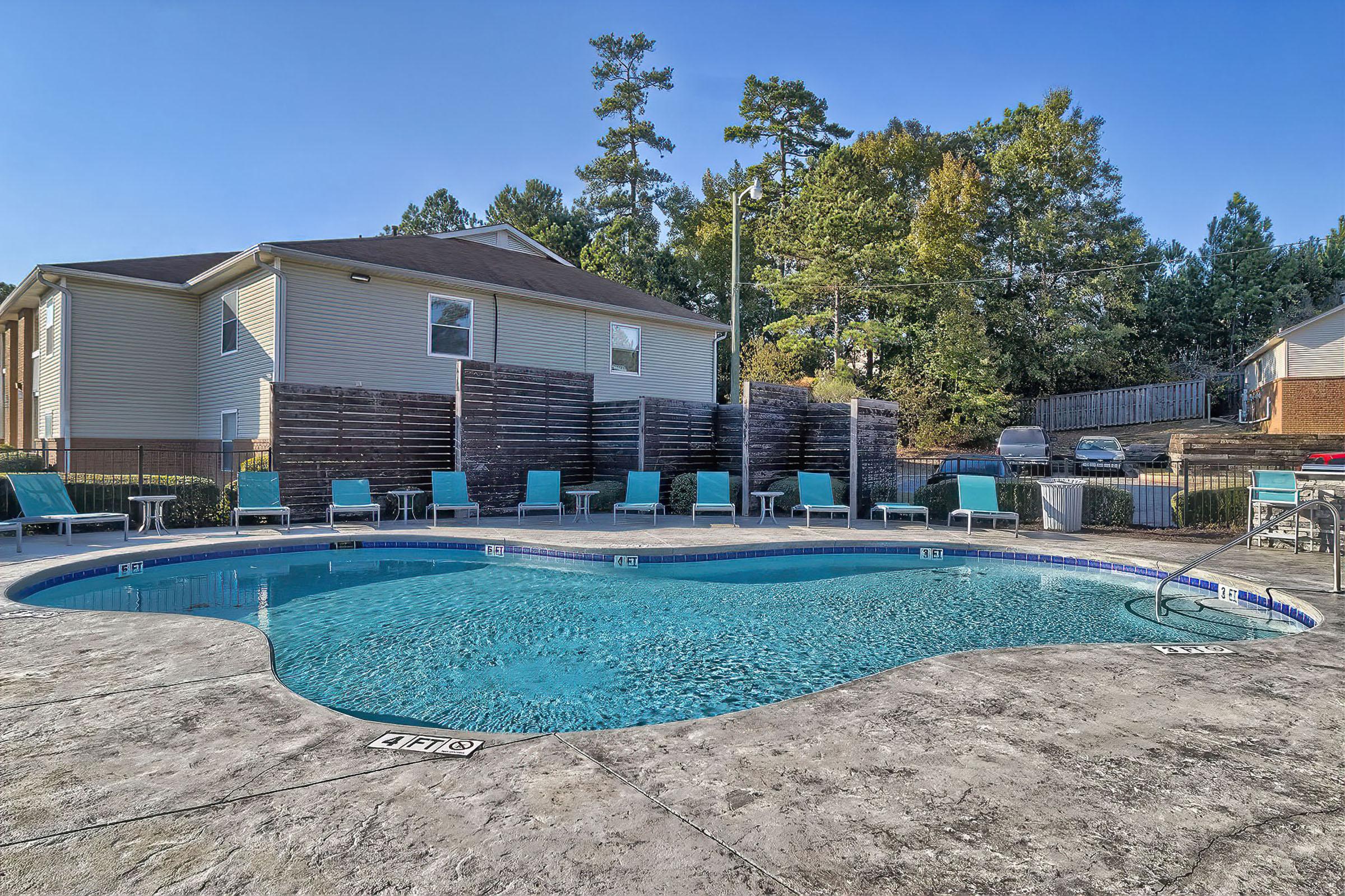 A sparkling outdoor swimming pool surrounded by lounge chairs in a sunny area. In the background, there is a two-story building and trees lining the property. The pool area features a textured concrete surface and is well-maintained, providing a relaxing atmosphere.