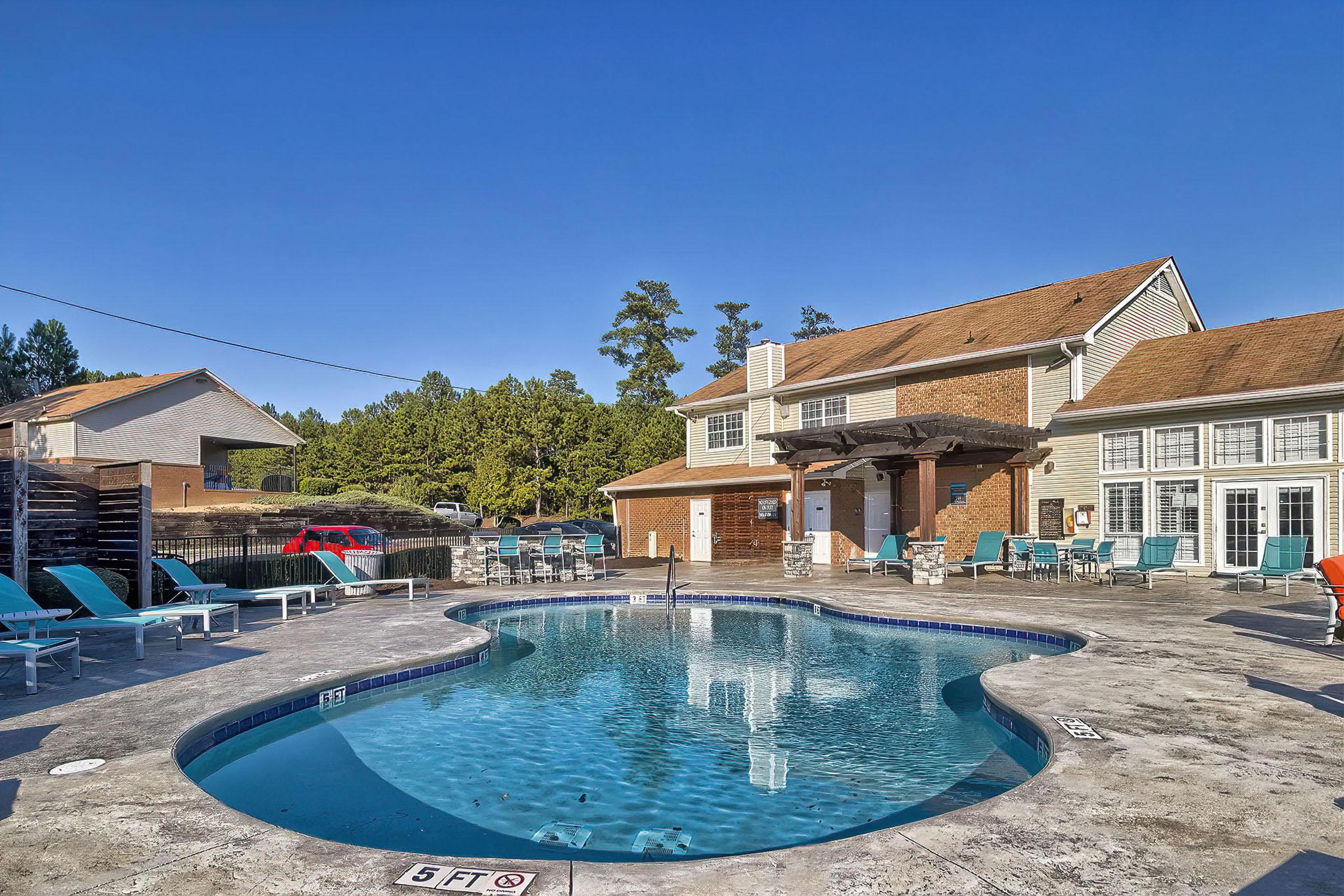 A clear blue swimming pool surrounded by lounge chairs and tables, with a two-story building in the background. Pine trees line the area, and the sky is bright and clear. The pool area features a depth marker, indicating a 5-foot depth.