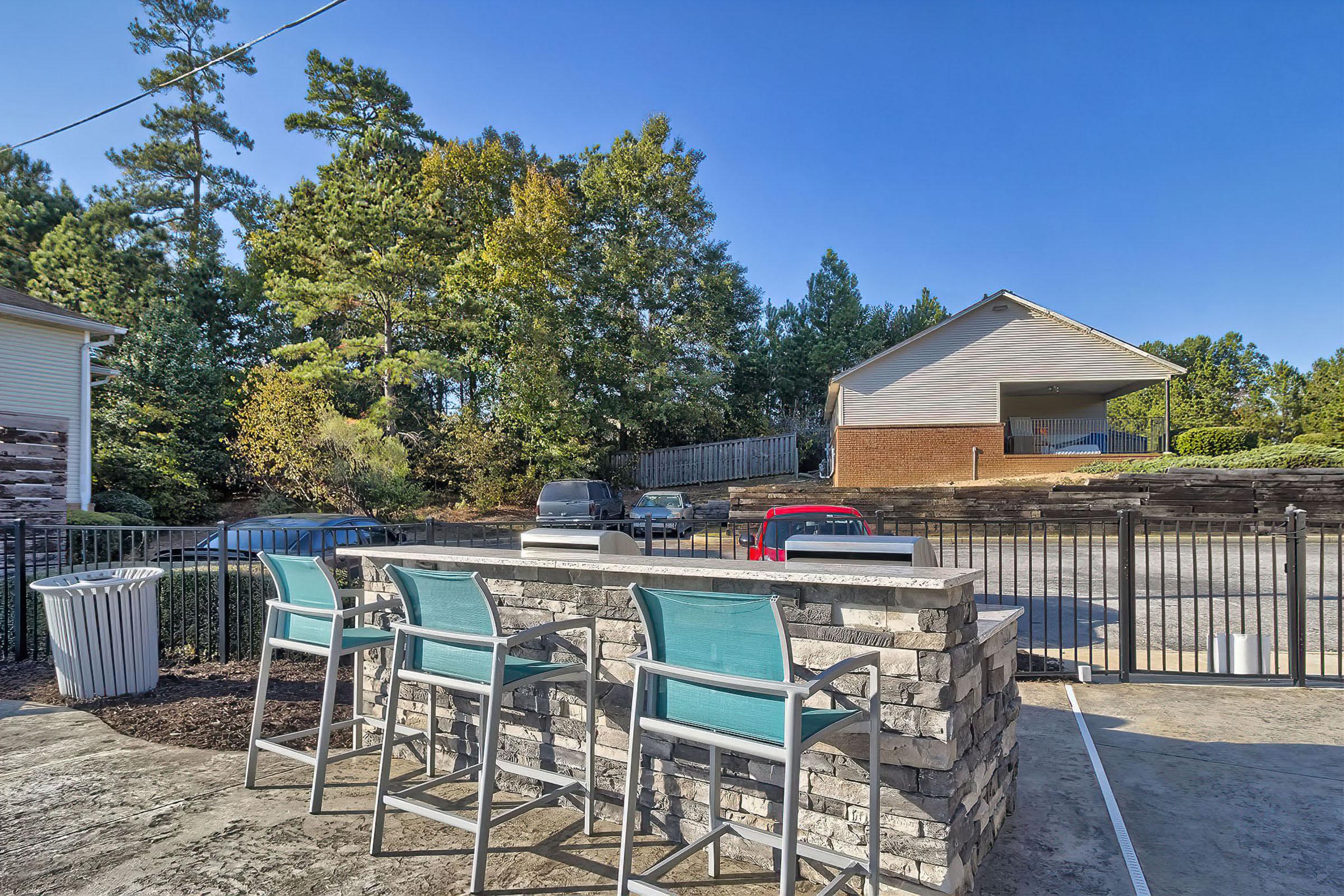 Outdoor space featuring a stone bar with turquoise high chairs, surrounded by trees. In the background, a house is visible, along with parked cars and a clean concrete patio. The sky is clear, indicating a bright and sunny day.