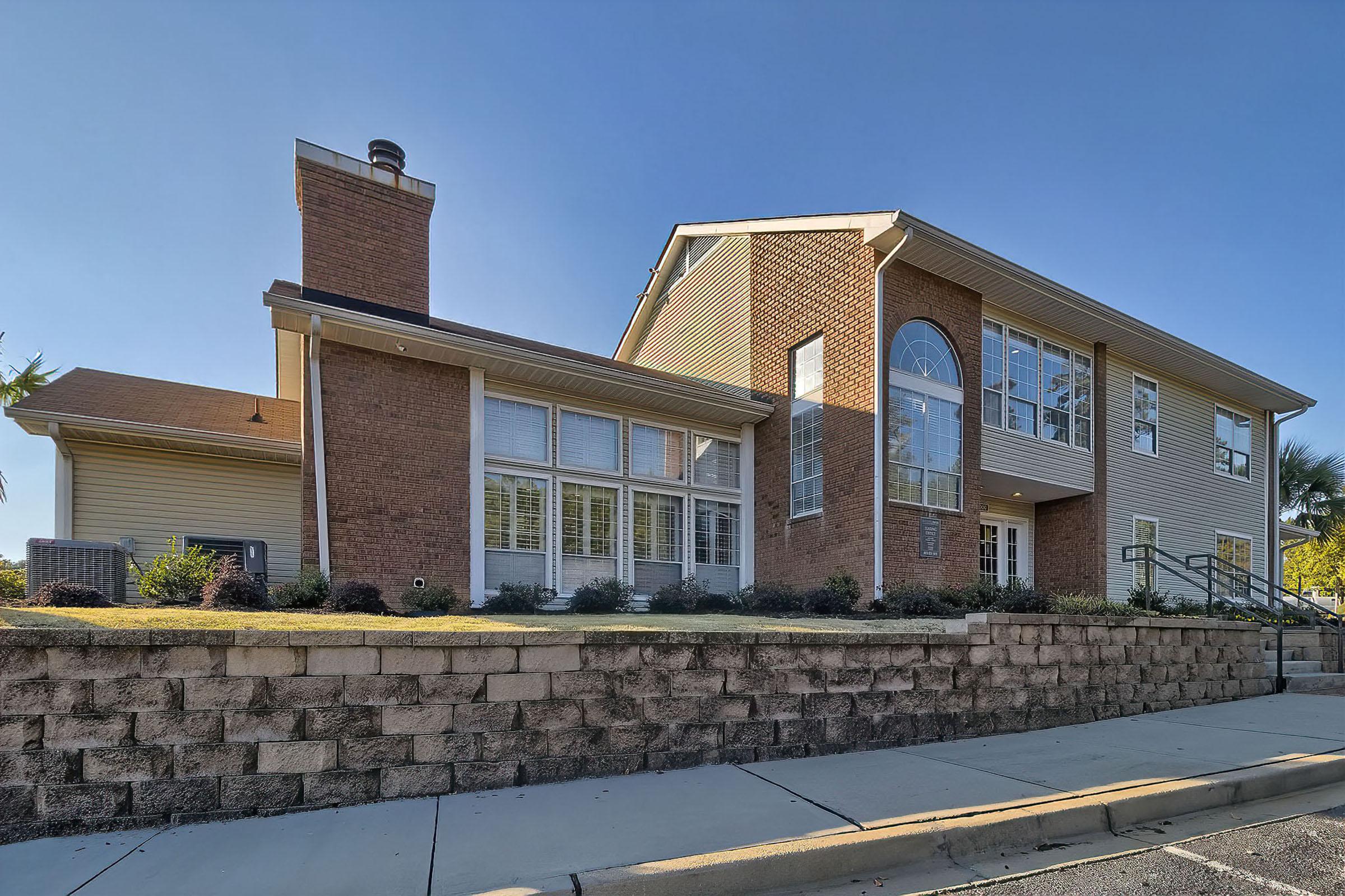 A modern two-story building featuring a combination of brick and siding exteriors. Large windows allow for natural light, and the entryway has a prominent arch. The structure is surrounded by landscaped greenery and a stone wall in front, set against a clear blue sky.