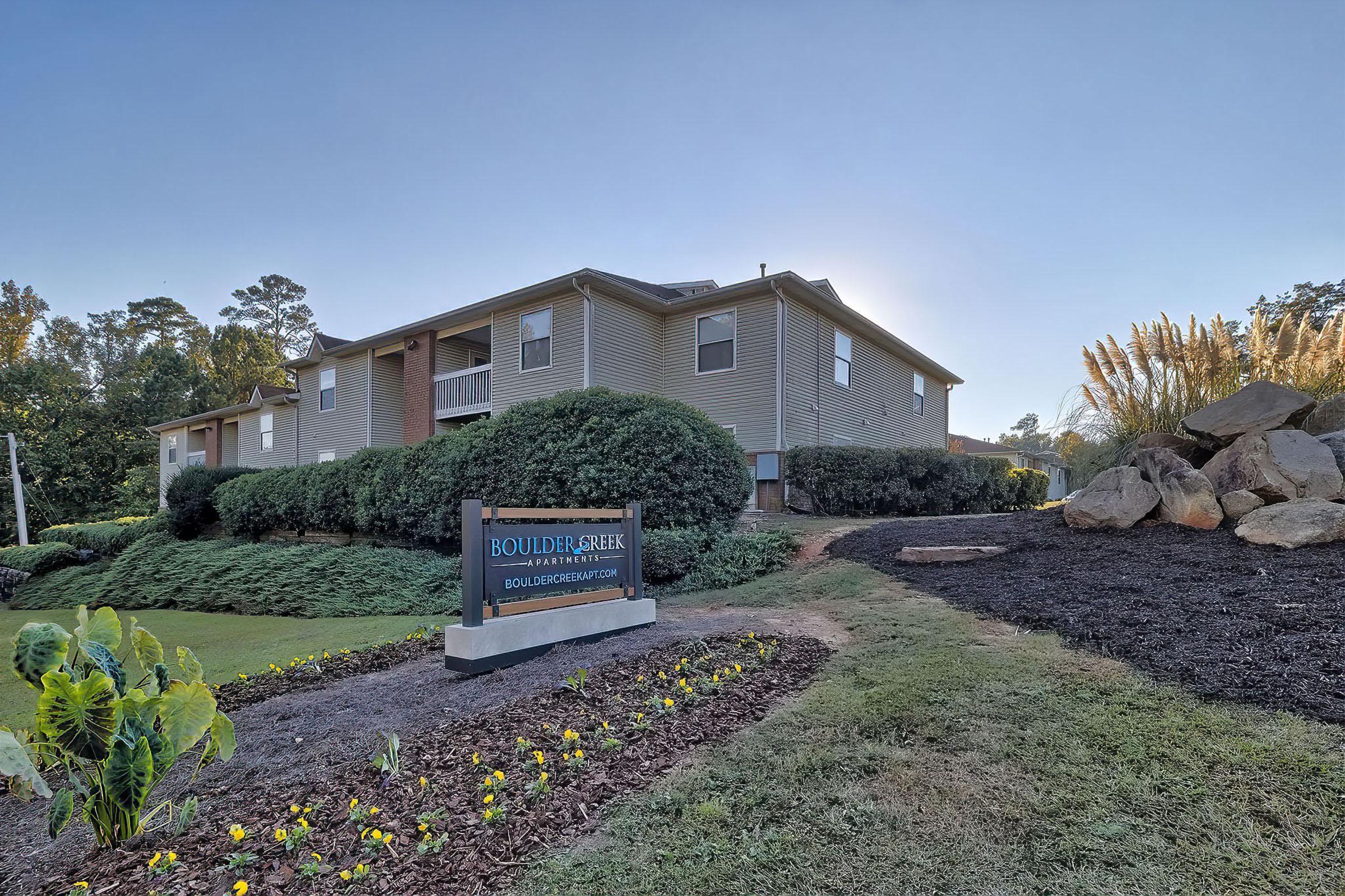 A view of Boulder Creek residential complex featuring a two-story building surrounded by well-maintained landscaping, including shrubs and flower beds. A sign with the name "Boulder Creek" is prominently displayed in front, along with a website URL. Clear skies and natural light enhance the scene.