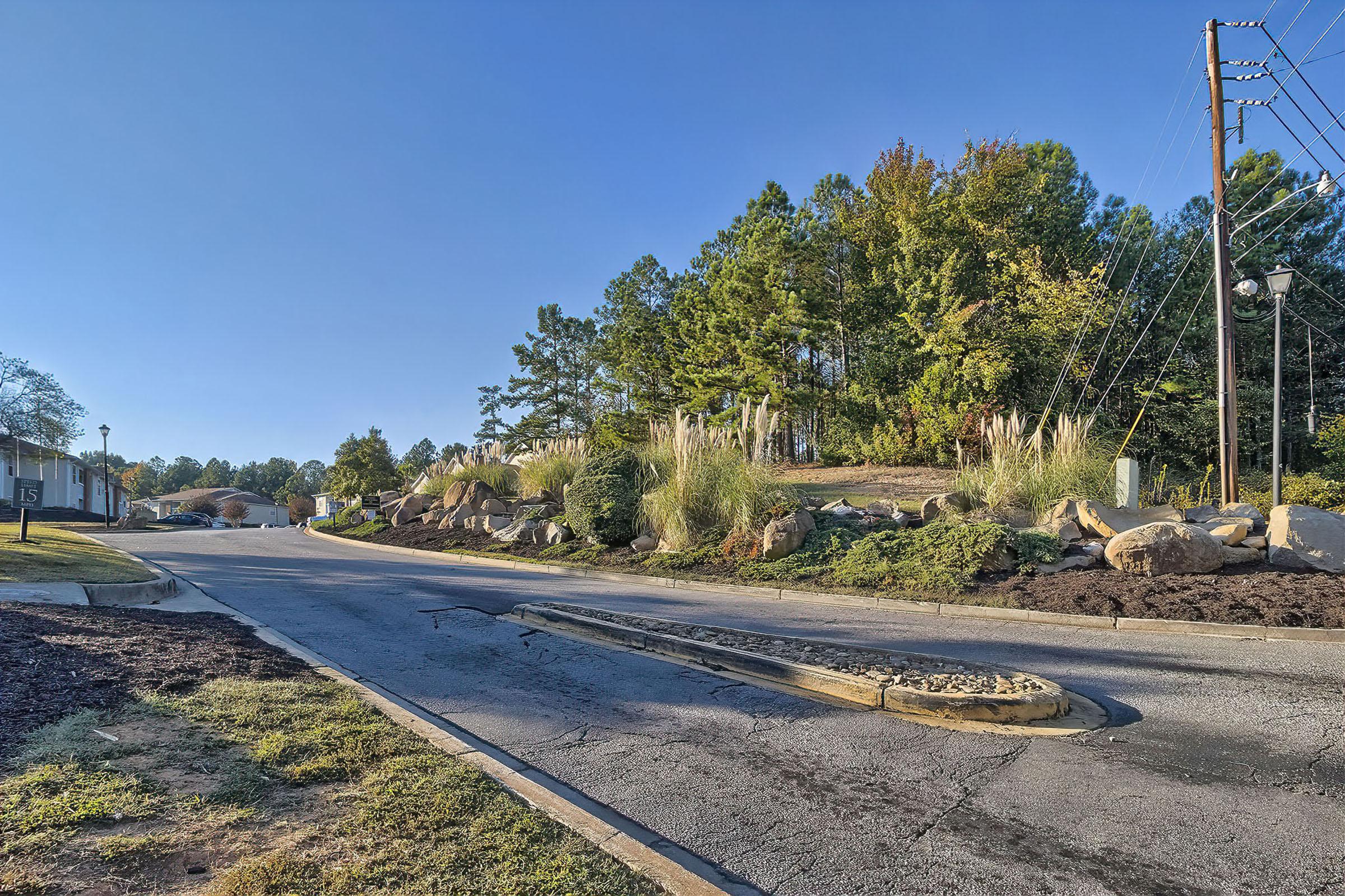 A view of a quiet street lined with trees and shrubs. In the foreground, a sloped roadway leads into a larger area, with rocks and vegetation visible. The sky is clear and blue, indicating a sunny day. Utility poles are seen on the side, suggesting a residential or commercial zone nearby.