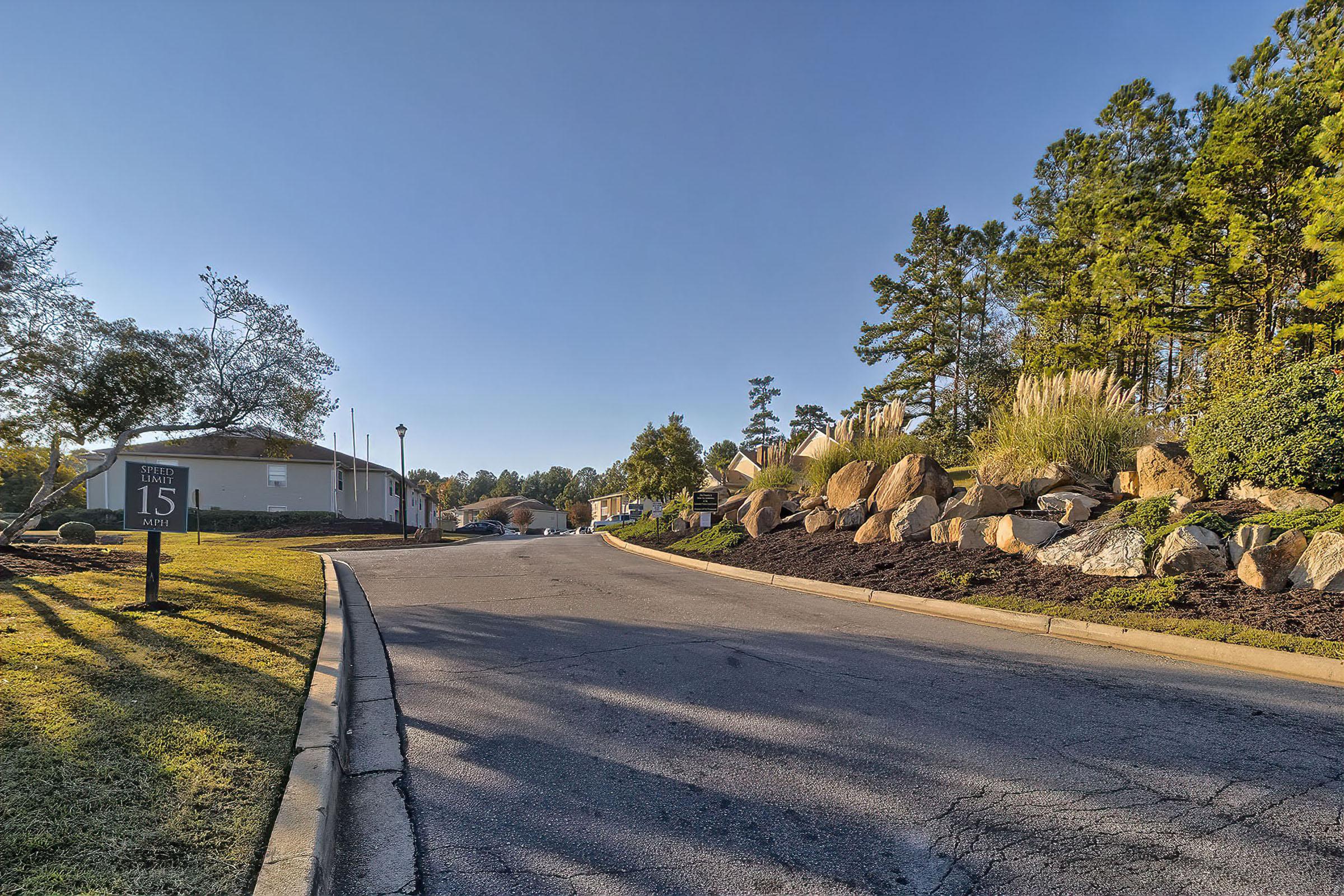 A peaceful neighborhood scene showing a winding road leading toward residential buildings. Lush greenery and large rocks line the roadside, with trees in the background. A sign indicating "Street 15" is visible on the left, under clear blue skies. The setting conveys a tranquil, suburban atmosphere.