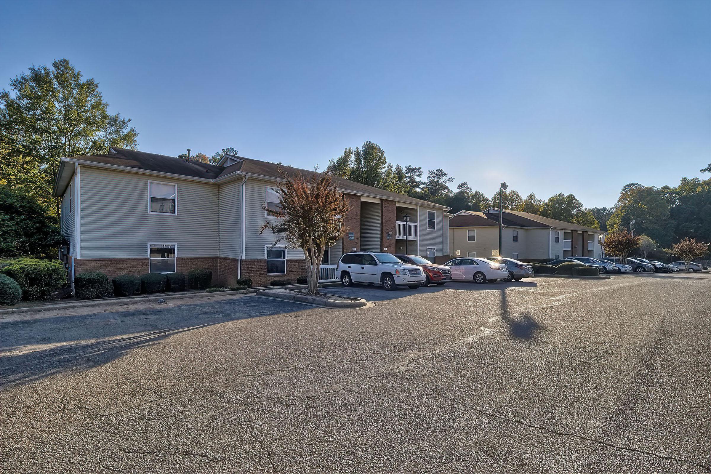 A low-rise apartment community with two buildings side by side, featuring a parking lot filled with vehicles. The scene is set during a clear, sunny day with trees and shrubs lining the property, creating a welcoming residential environment.