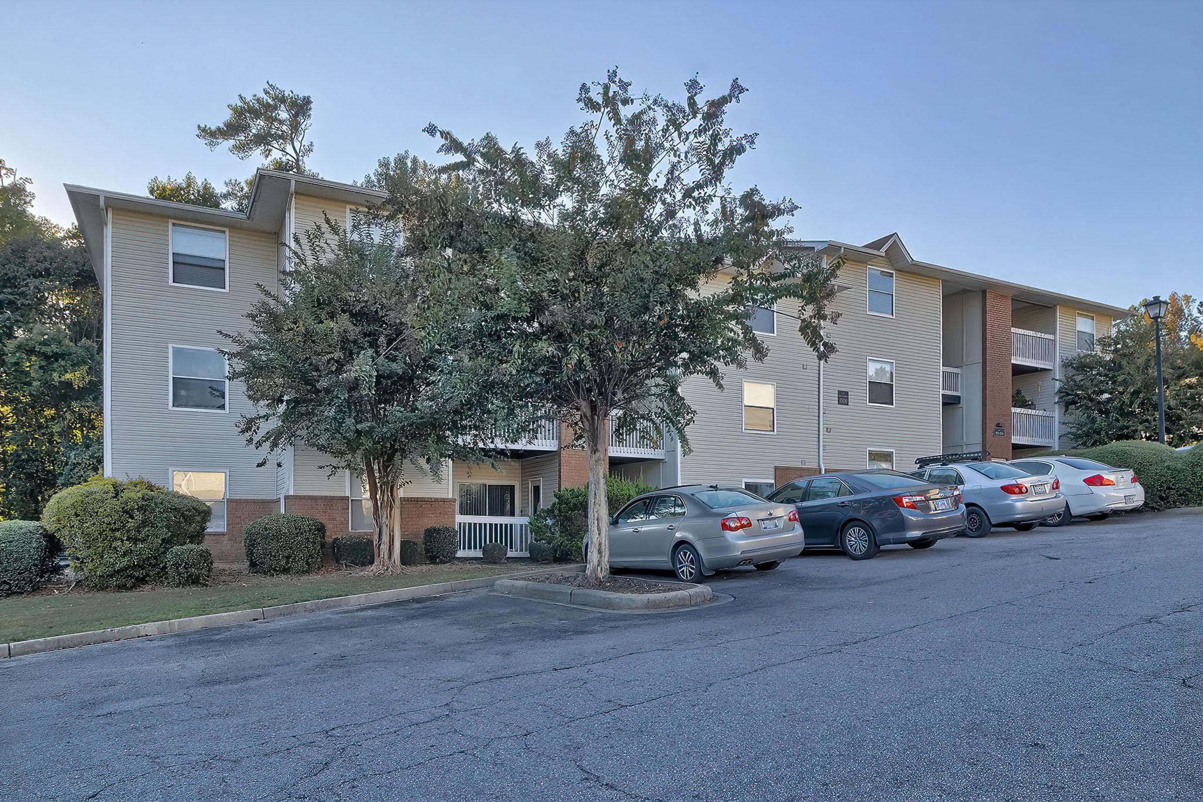 A three-story residential building surrounded by trees and shrubs, with a parking lot in front. Several cars are parked along the drive, and the building features a mixture of windows and balconies. The setting is a suburban area under a clear sky.