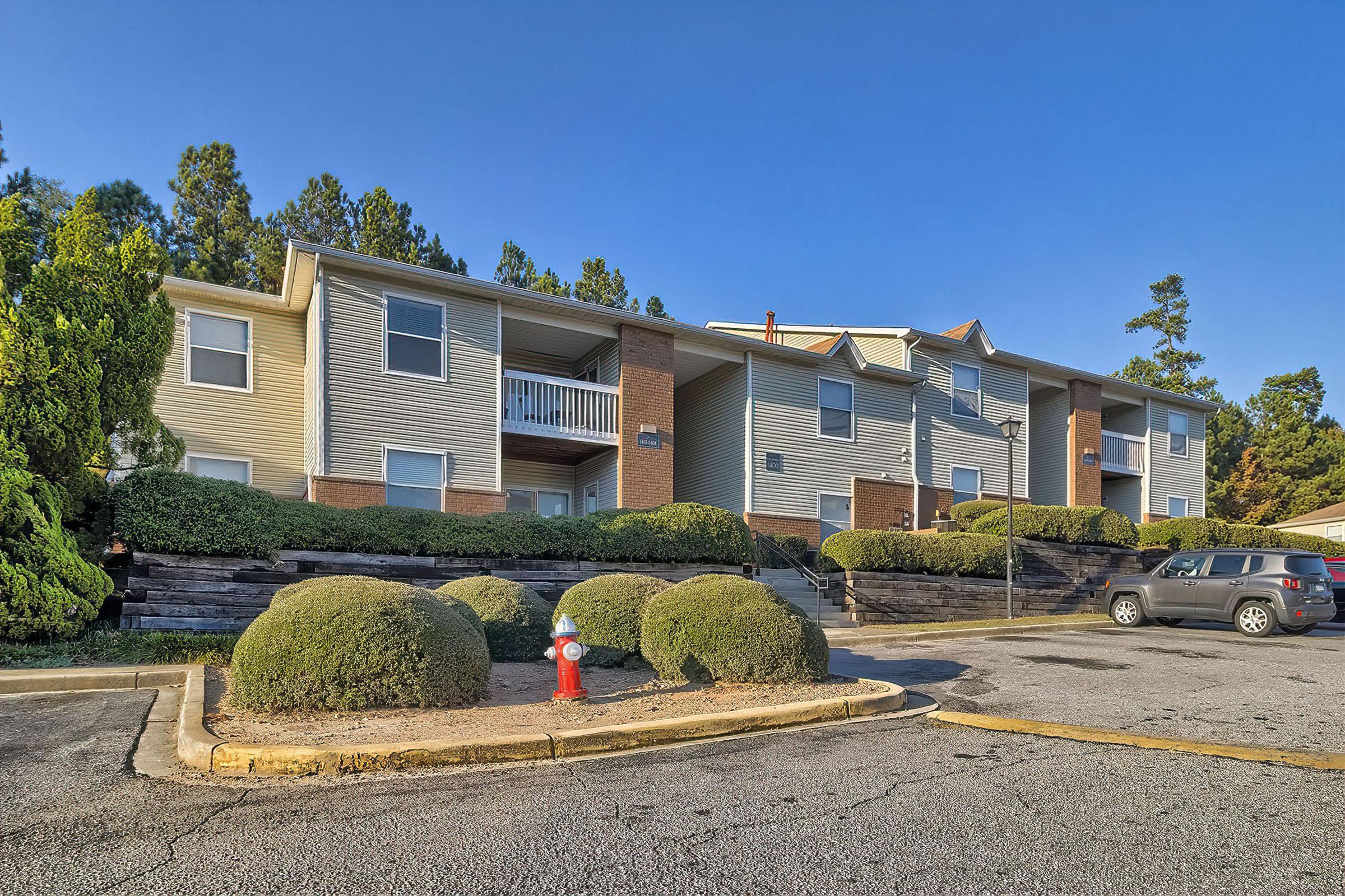 A multi-story apartment building surrounded by neatly trimmed shrubs and bushes. A fire hydrant is visible in the foreground, with a paved parking lot. Trees are visible in the background under a clear blue sky.