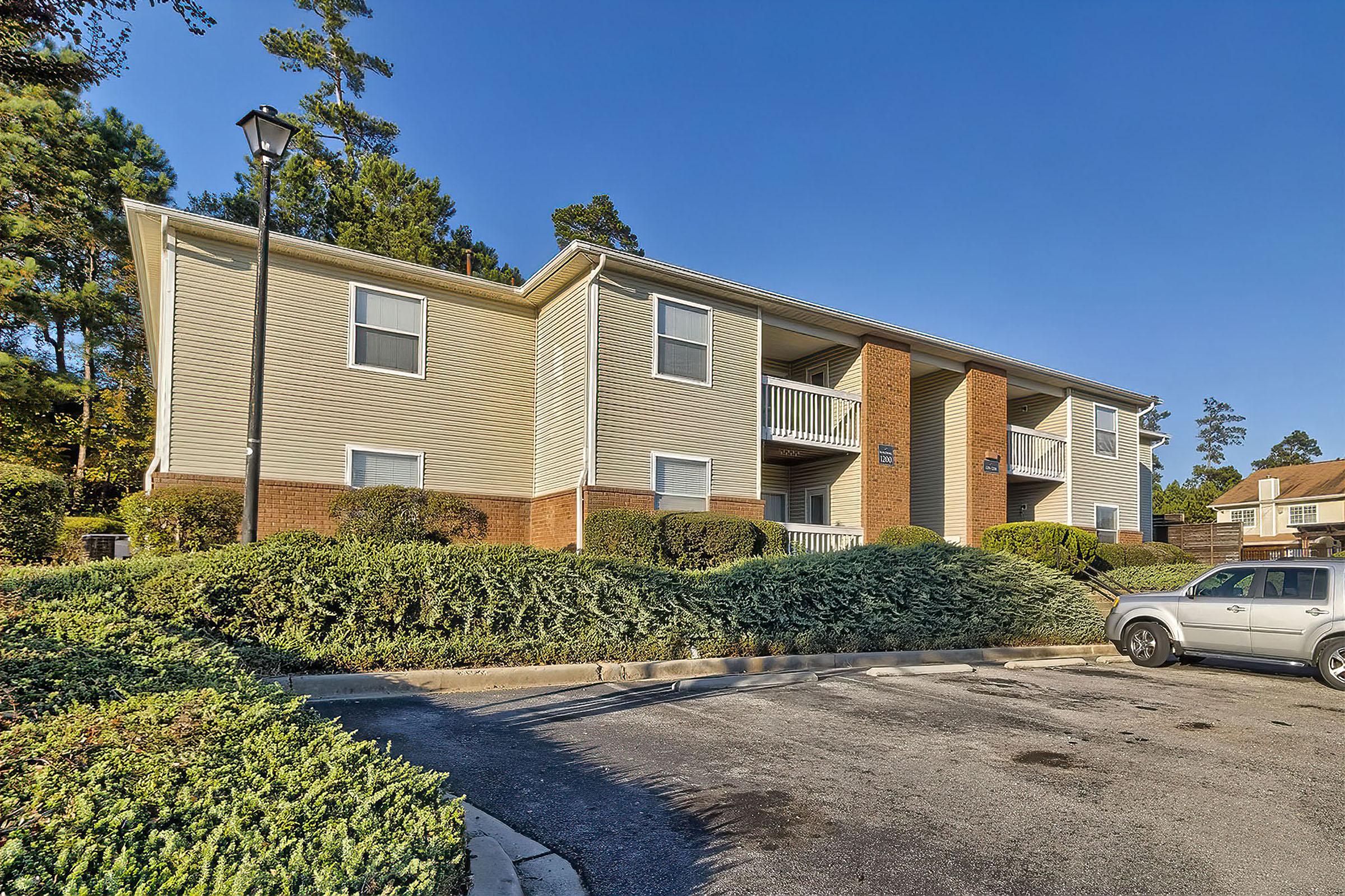 A two-story apartment building with beige siding and brick accents, surrounded by neatly trimmed bushes and shrubs. A parking area is visible in front, with a white car parked nearby. Trees and clear blue skies are in the background, creating a serene residential atmosphere.