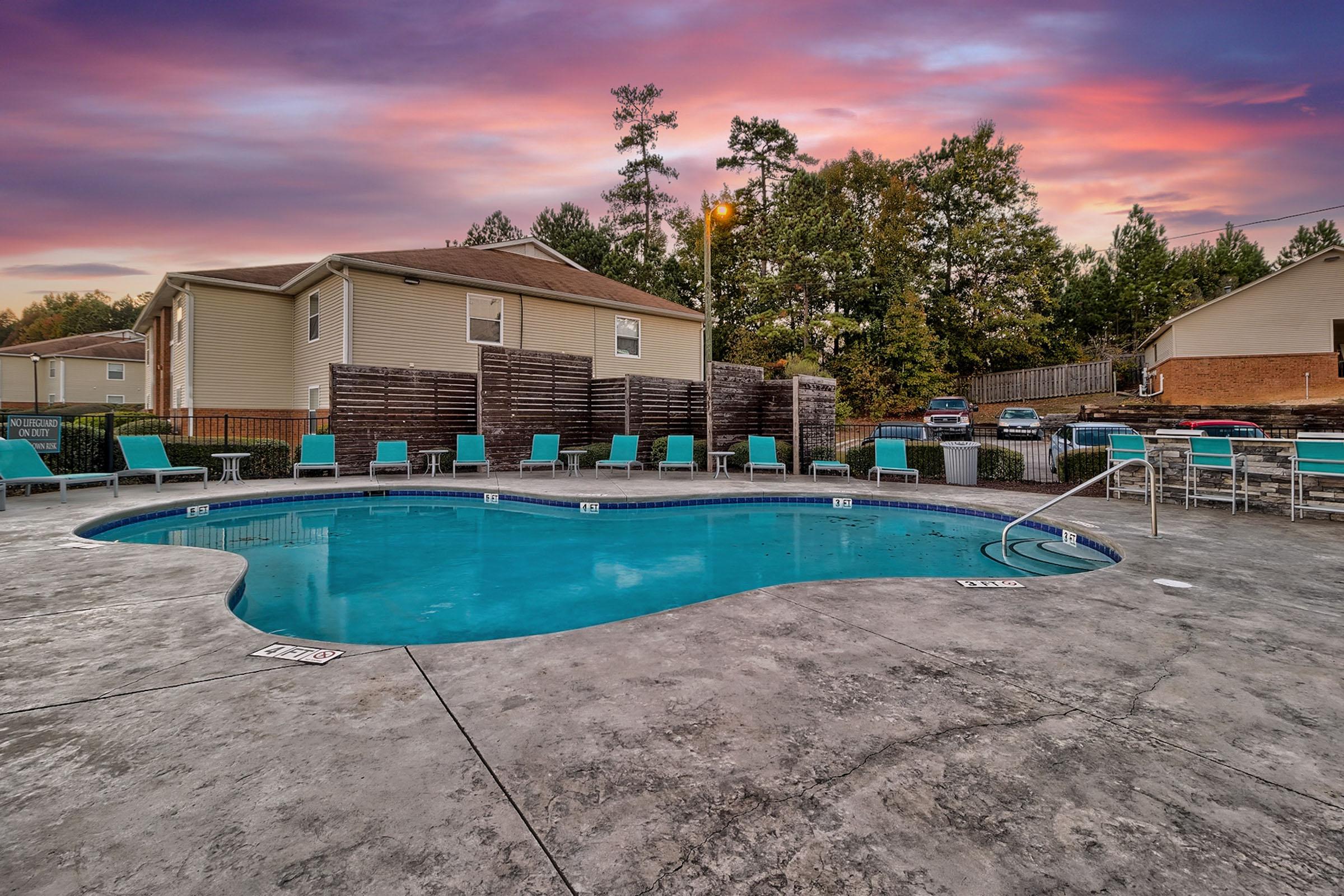 A beautifully lit outdoor swimming pool surrounded by lounge chairs, set against a vibrant sunset with pink and purple hues. Trees and a few residential buildings are visible in the background, creating a relaxing atmosphere. The pool area is designed for leisure and enjoyment.