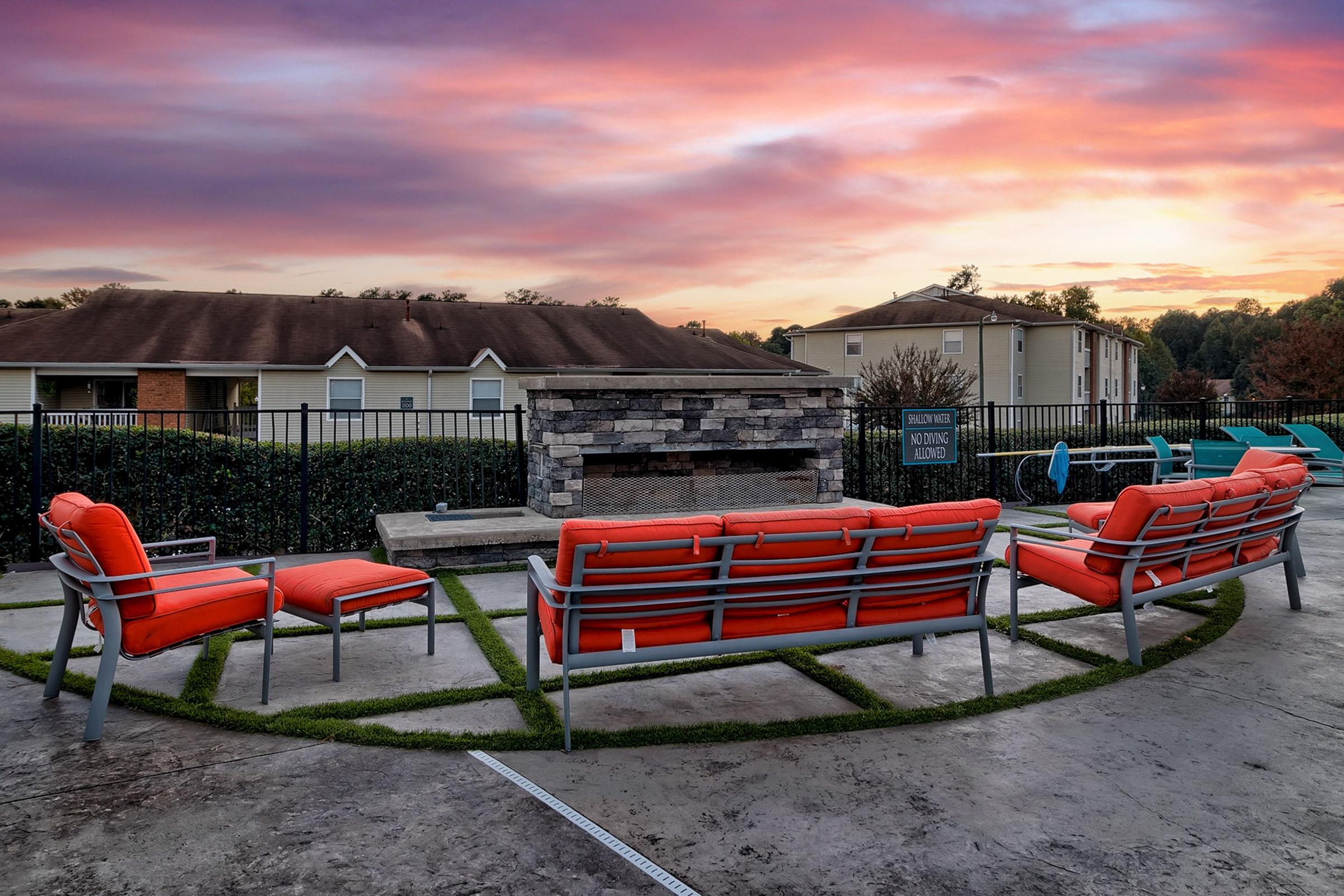 An outdoor seating area featuring orange chairs arranged in a semi-circle around a stone fireplace, with a backdrop of colorful sunset skies. The setting is surrounded by green landscaping and appears to be part of a residential complex.