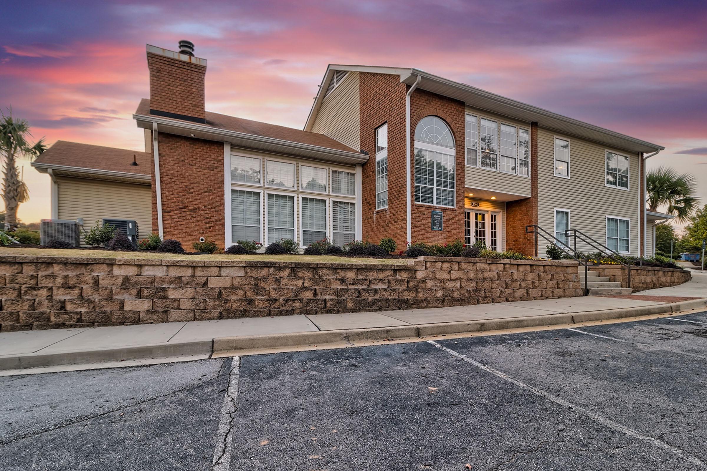 A modern two-story brick and siding building with large front windows, surrounded by landscaped gardens. The sky features a vibrant sunset with shades of purple and orange. A stone wall borders the parking area, which has clearly defined spaces and is empty.